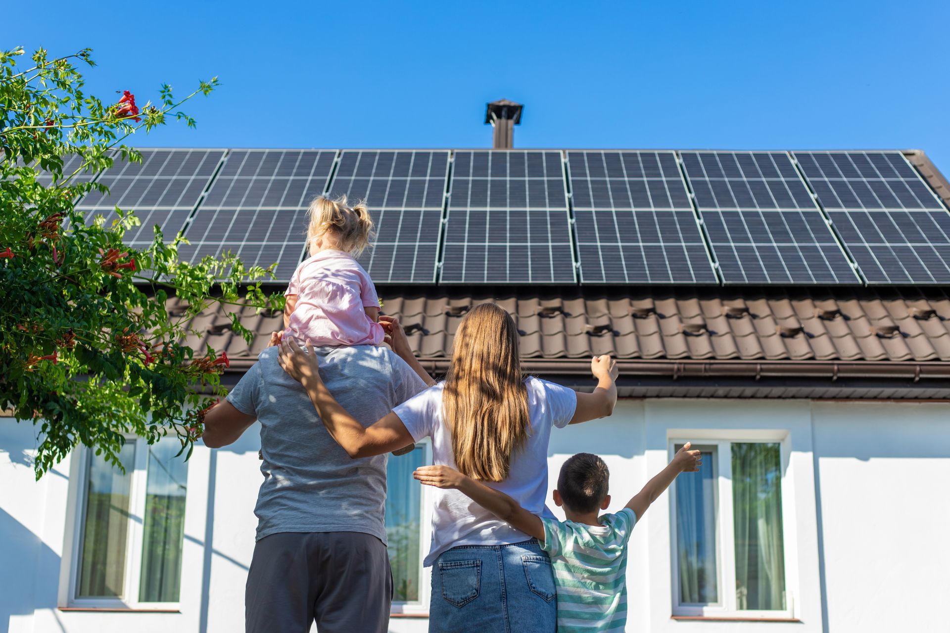 Family standing outside a house, looking up at rooftop solar panels under a clear blue sky