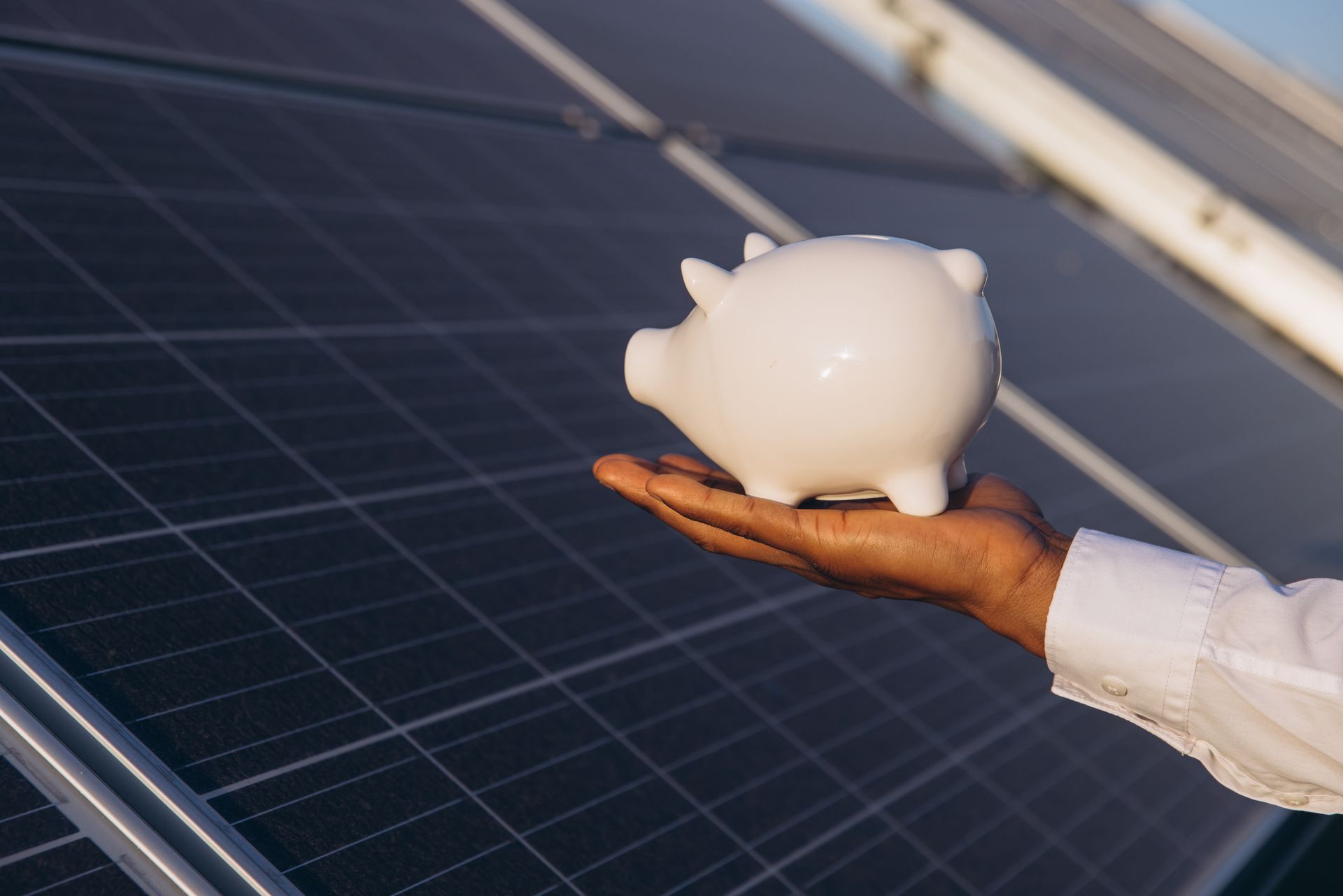 Hand holding a white piggy bank beside solar panels, symbolizing renewable energy savings
