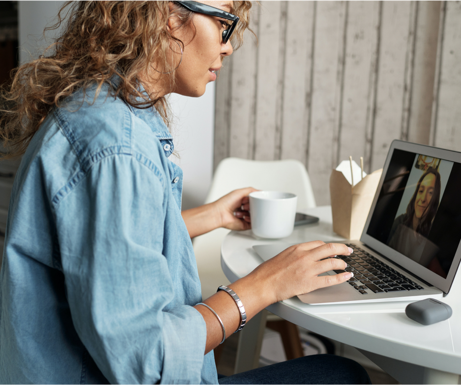 Woman typing on a laptop at a white desk, with a coffee cup and a video call on screen.