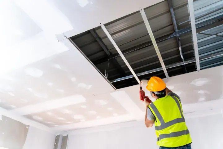 Worker in hard hat installing ceiling panels in a bright room with exposed ceiling tiles.