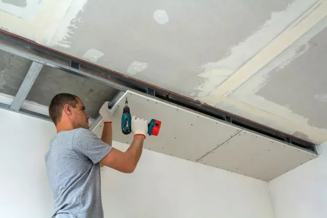 Worker installing drywall on a ceiling with a drill and red tool in an unfinished room