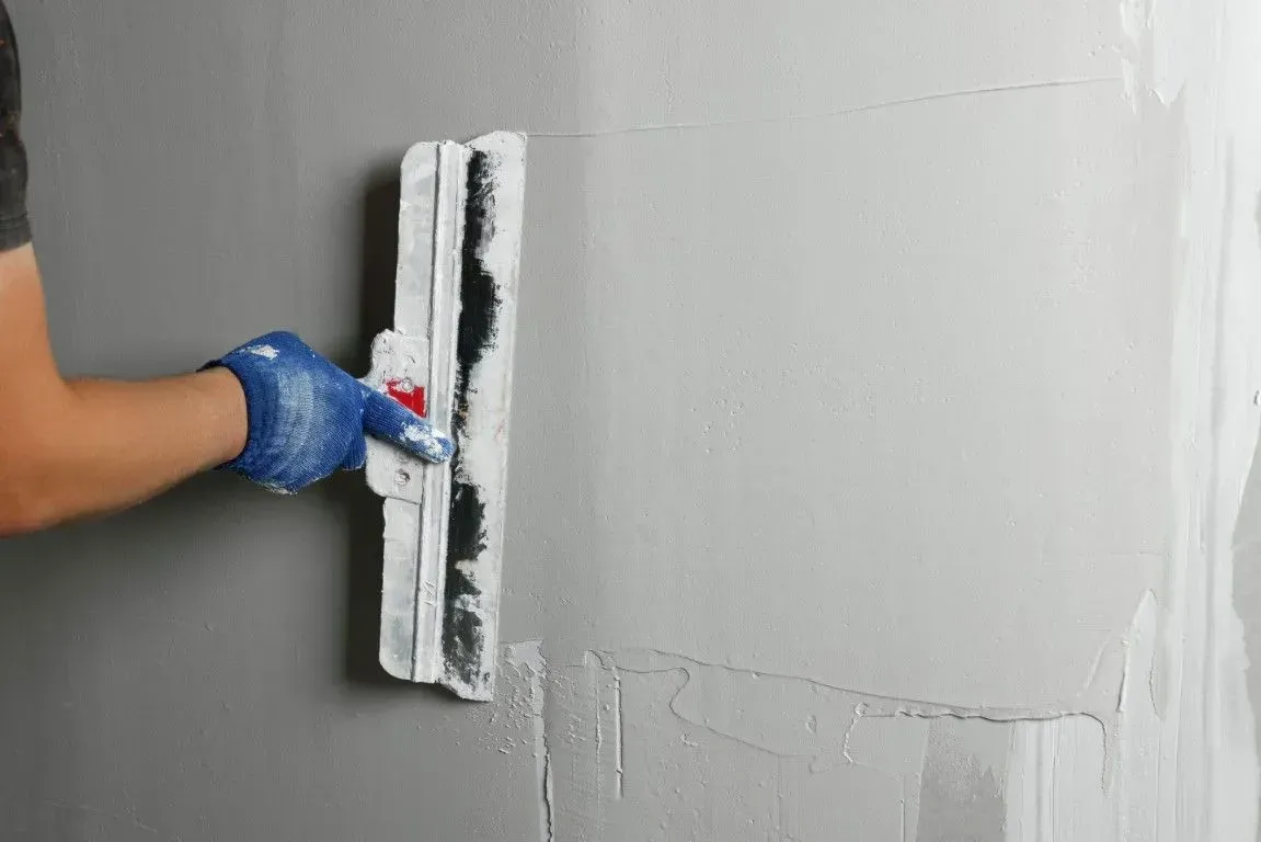 Gloved hand smoothing gray plaster on a wall with a trowel