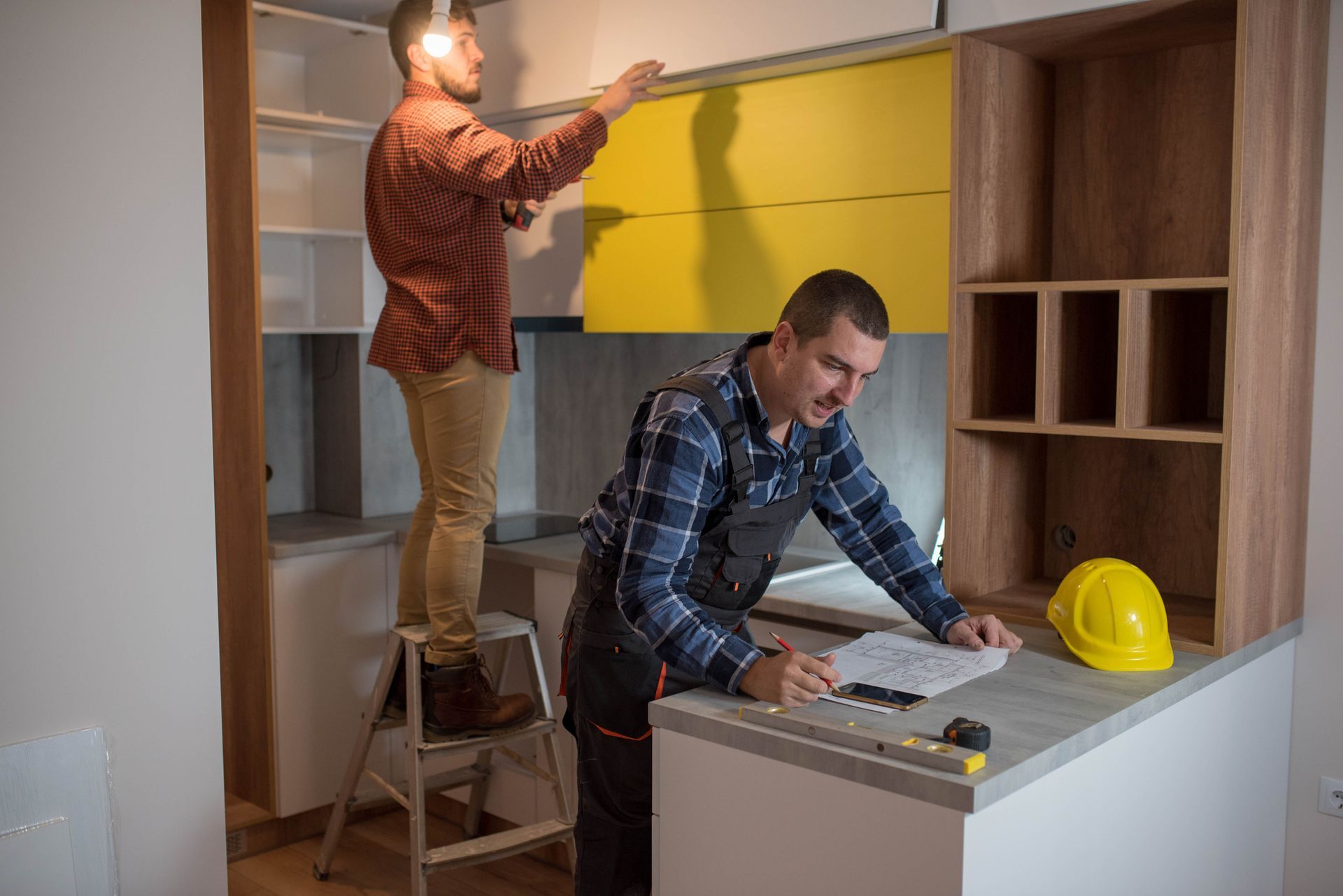 Two men are assembling a kitchen cabinet in a workshop as part of a kitchen remodeling project.