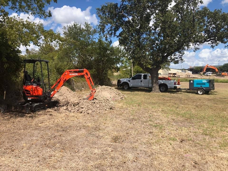 Orange excavator digging dirt, next to a white truck with a trailer, near trees and a building.