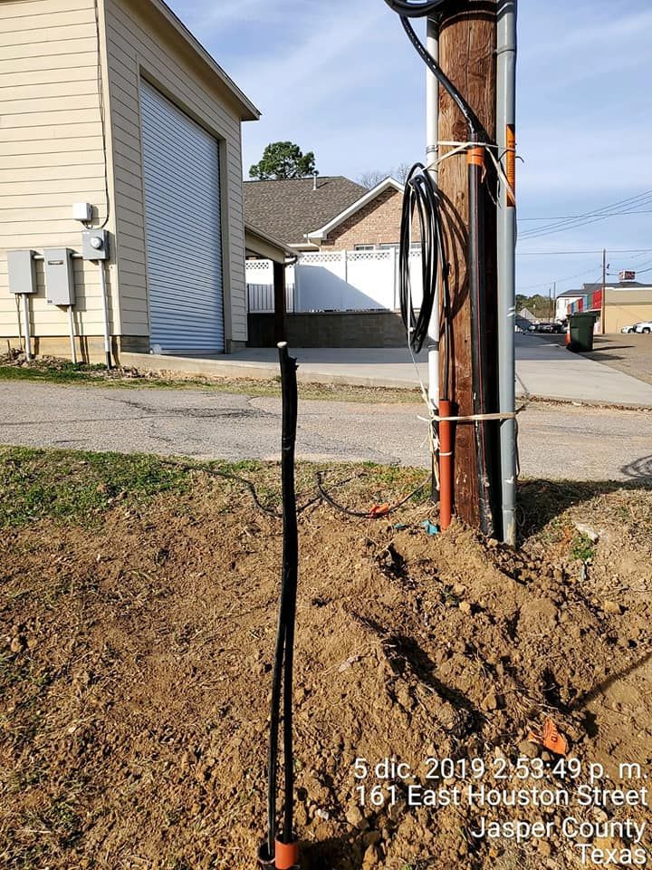 Black cable emerging from the ground near a utility pole with wires and conduits. A building is in the background.