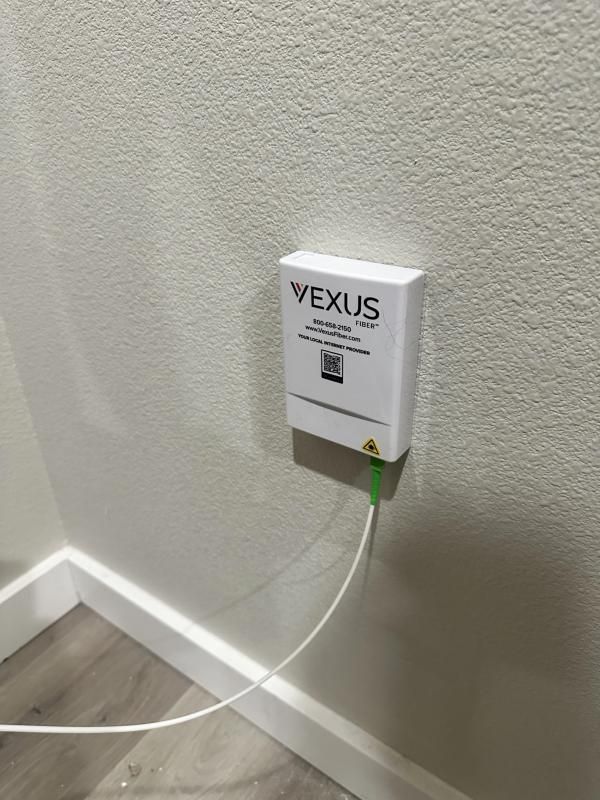 White Wexus fiber optic box mounted on a white wall, green fiber cable plugged in, next to a light wood-colored floor.