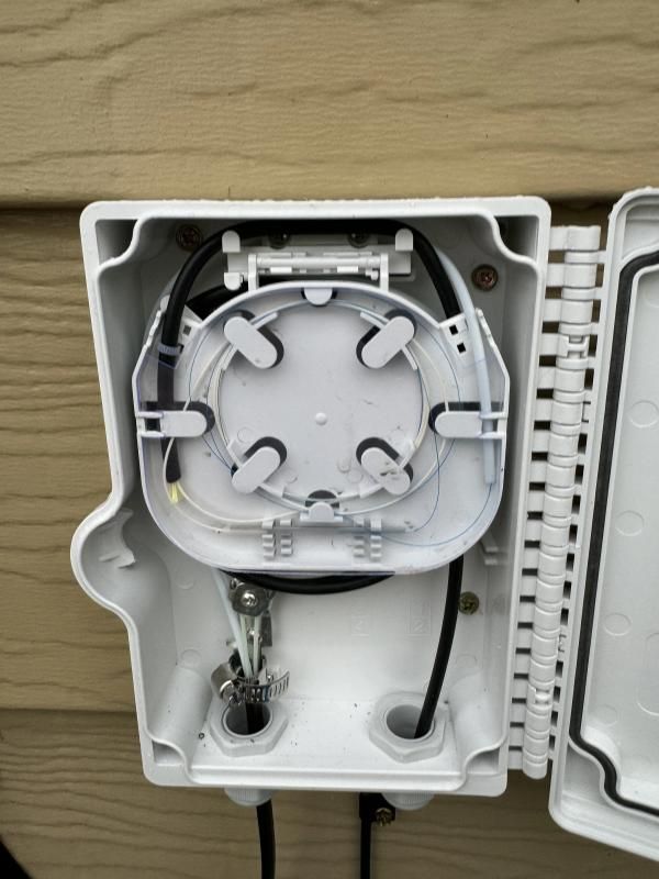 Open white outdoor fiber optic terminal box on a wooden wall. Black cables, white components.