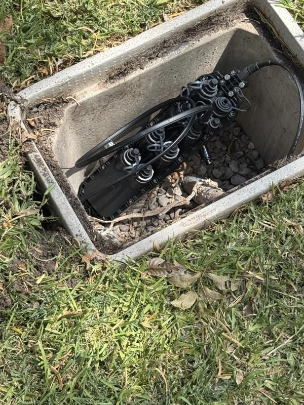 Open fiber optic cable junction box in a grassy yard. Black cables and connectors visible.