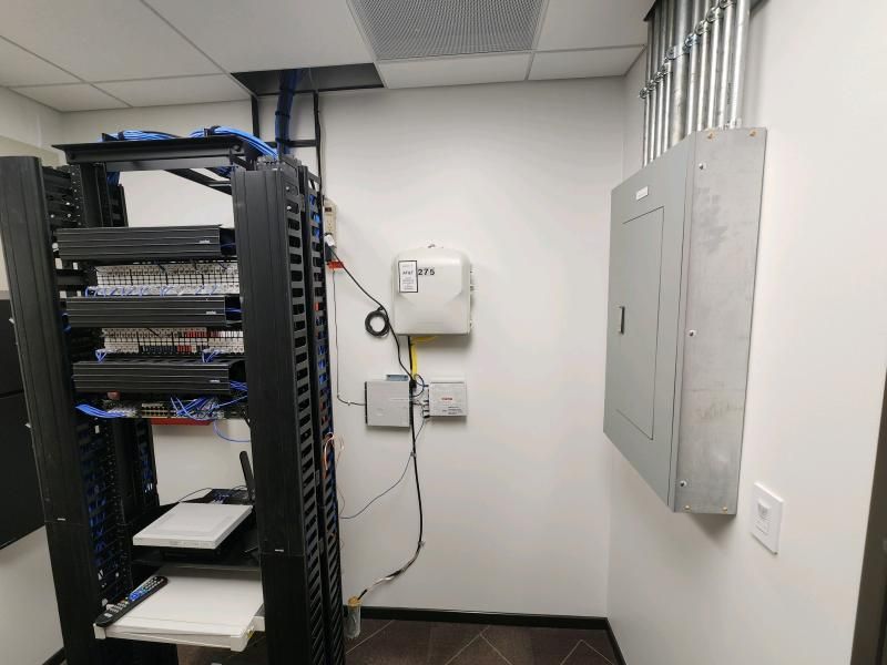 Server racks with network equipment and electrical panel in a room. Cables and wires are visible.