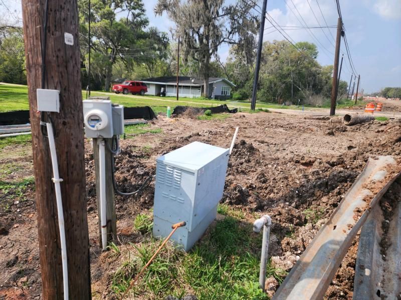 Utility equipment, including a transformer, meter, and wires, sits in a muddy area near a road and a house.