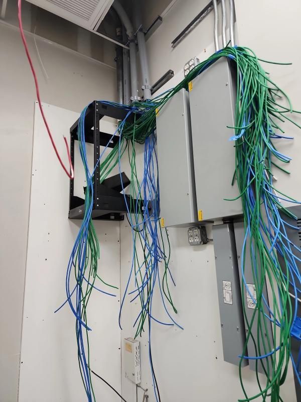 Network cables in a server room. Blue and green cables hang from a black rack and a gray box on a white wall.
