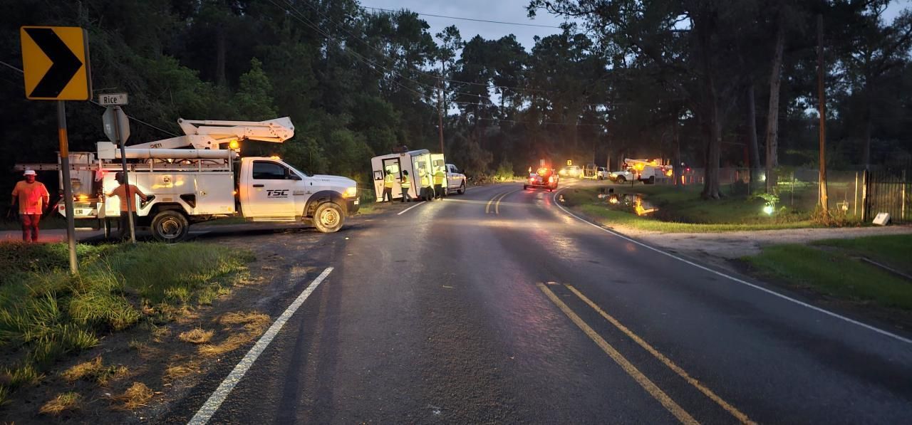 Utility trucks parked on a road at dusk; a worker stands by the road sign, and lights glow in the distance.