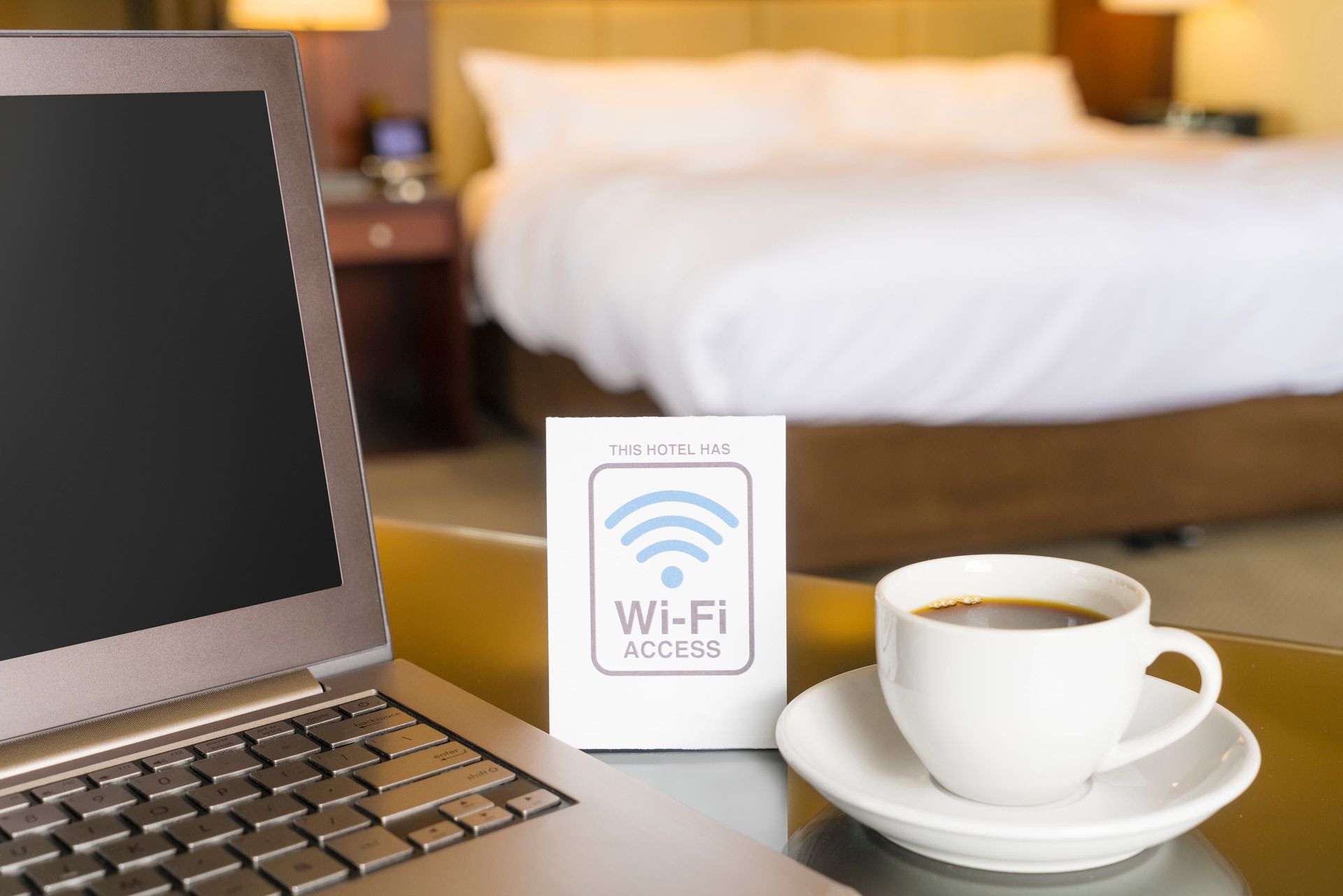 Laptop, Wi-Fi access box, and coffee cup on a table in a hotel room with a bed in the background.