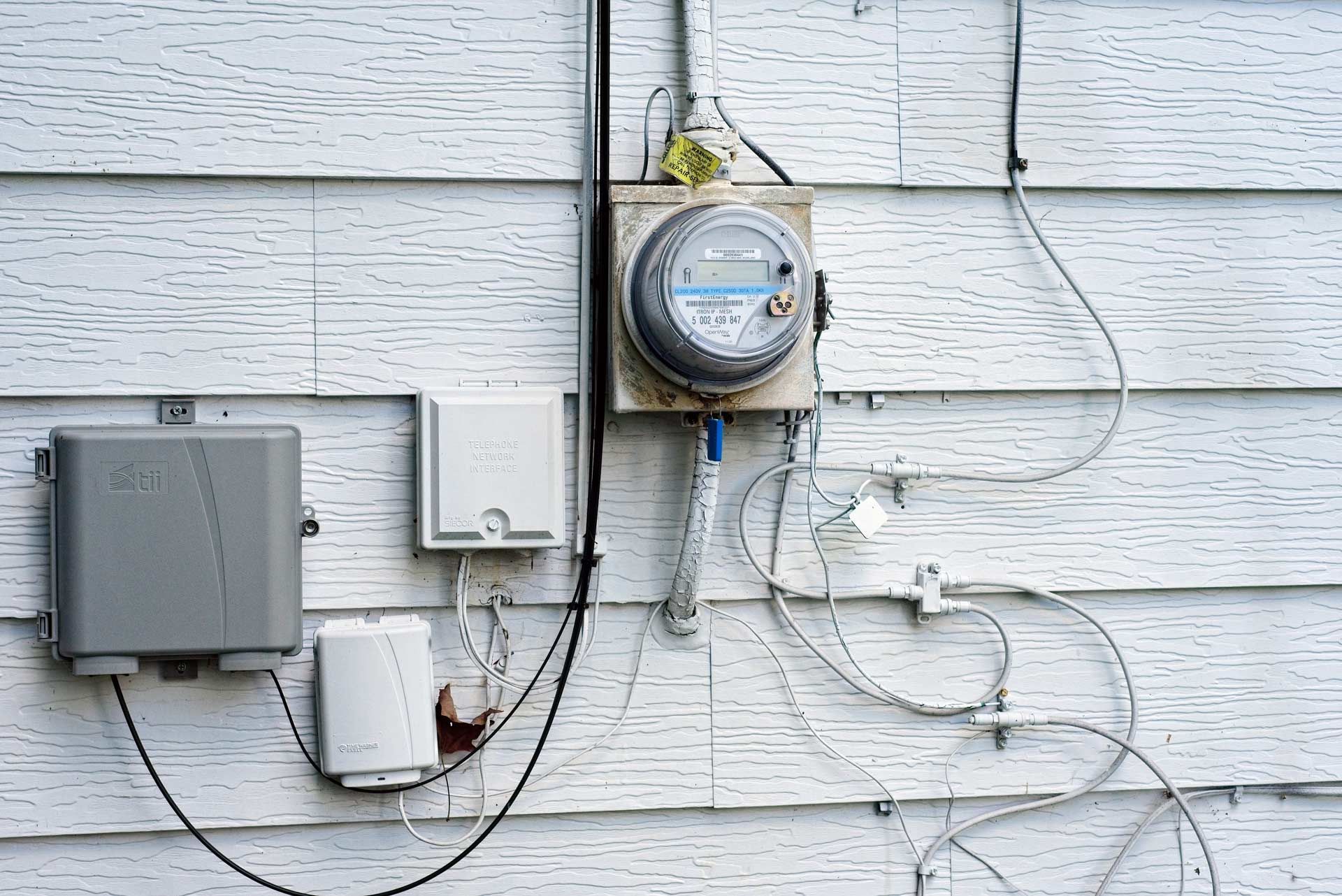 Electrical meter and various boxes on a white siding wall. Wires and cables are visible.