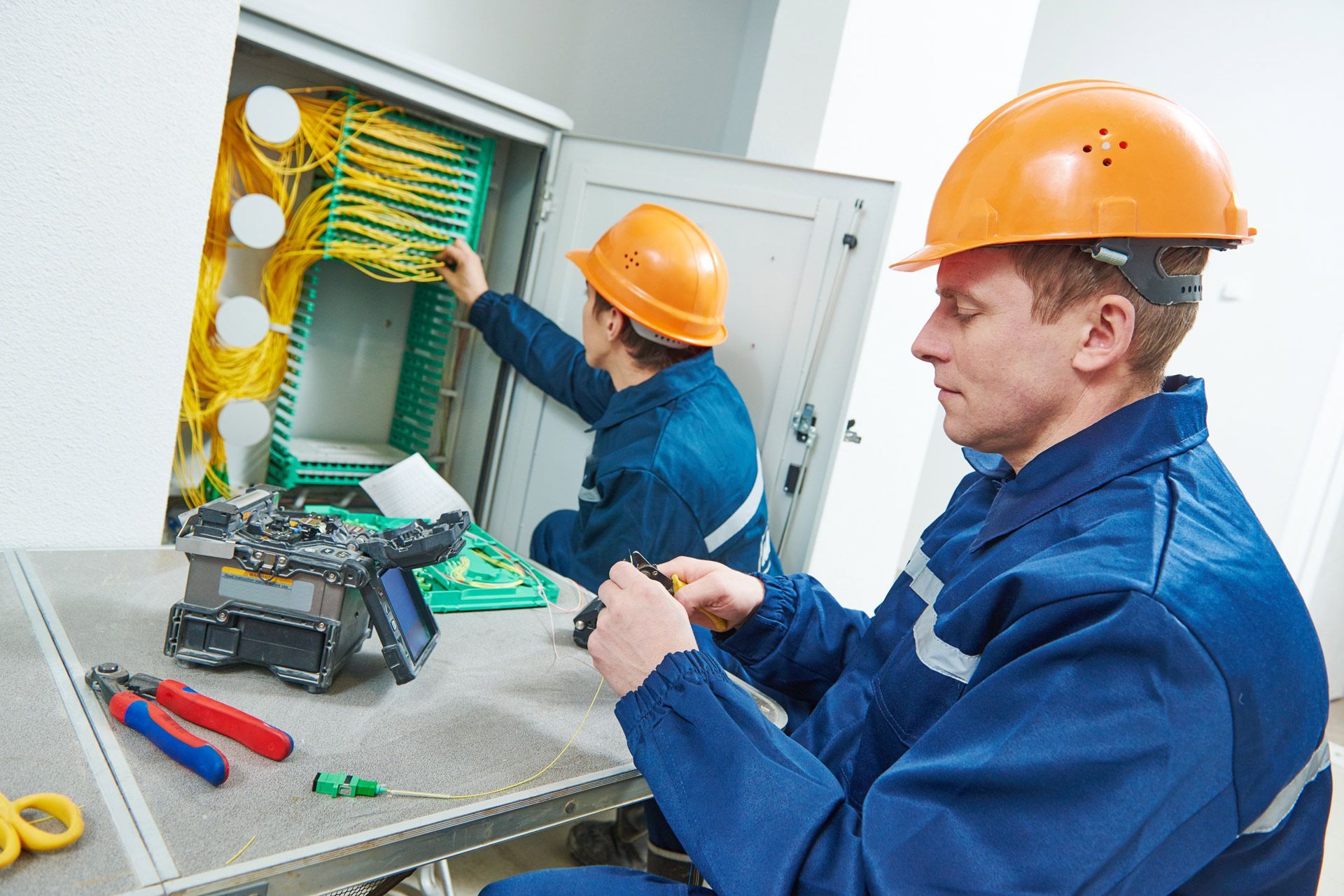 Two electricians in orange helmets and blue jumpsuits working on a network cable panel.