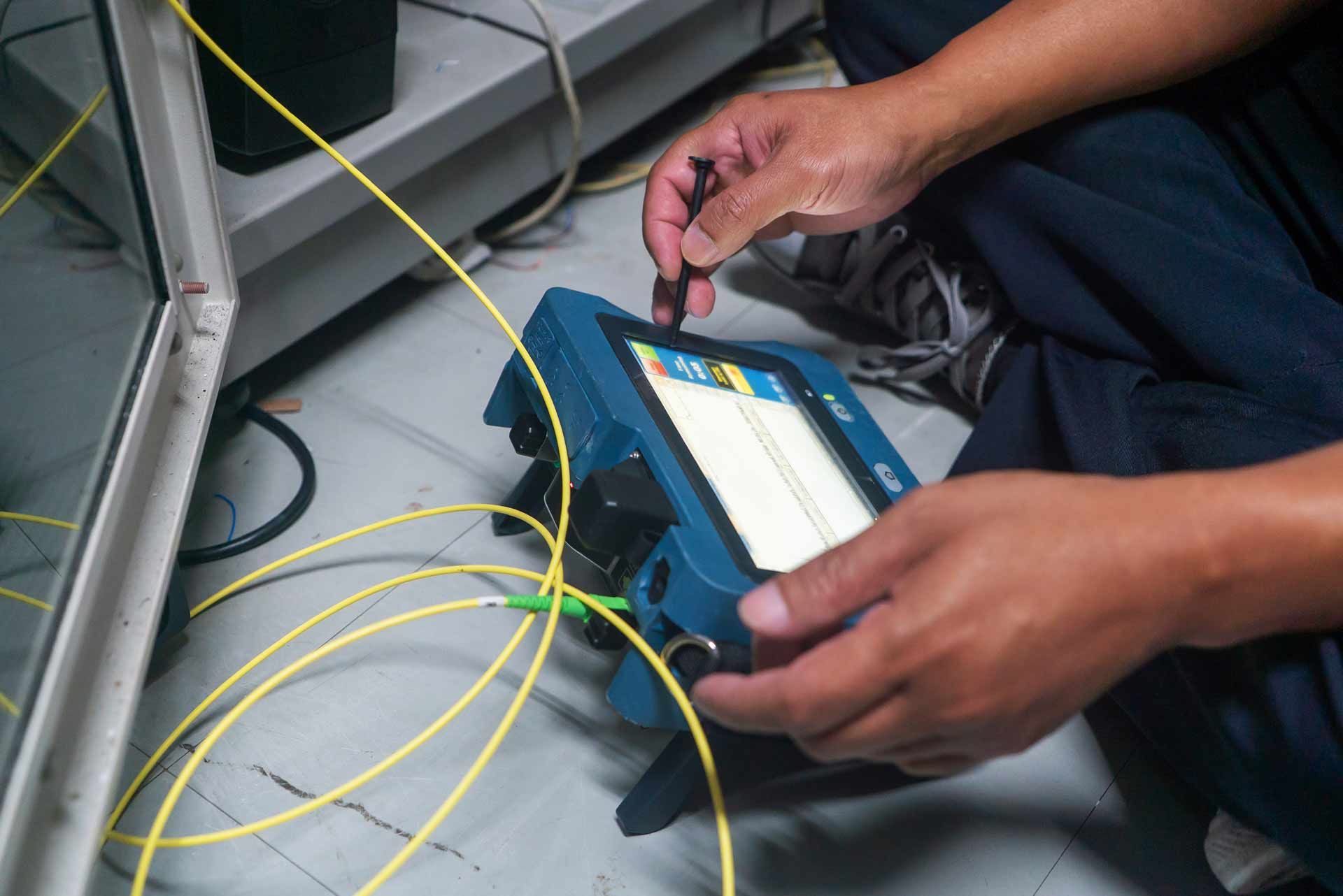 Person using a blue testing device, connected to yellow fiber optic cables, on a floor.