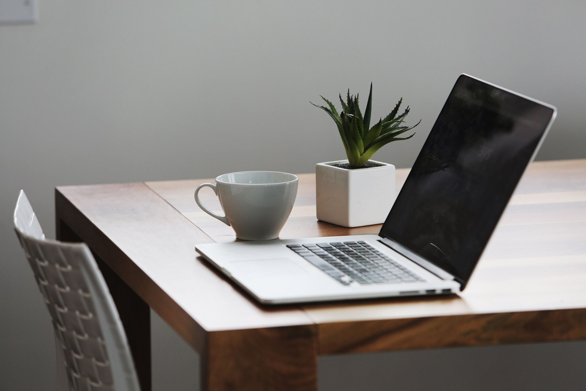 Laptop, mug, and plant on a wooden table, with a chair visible in the corner.