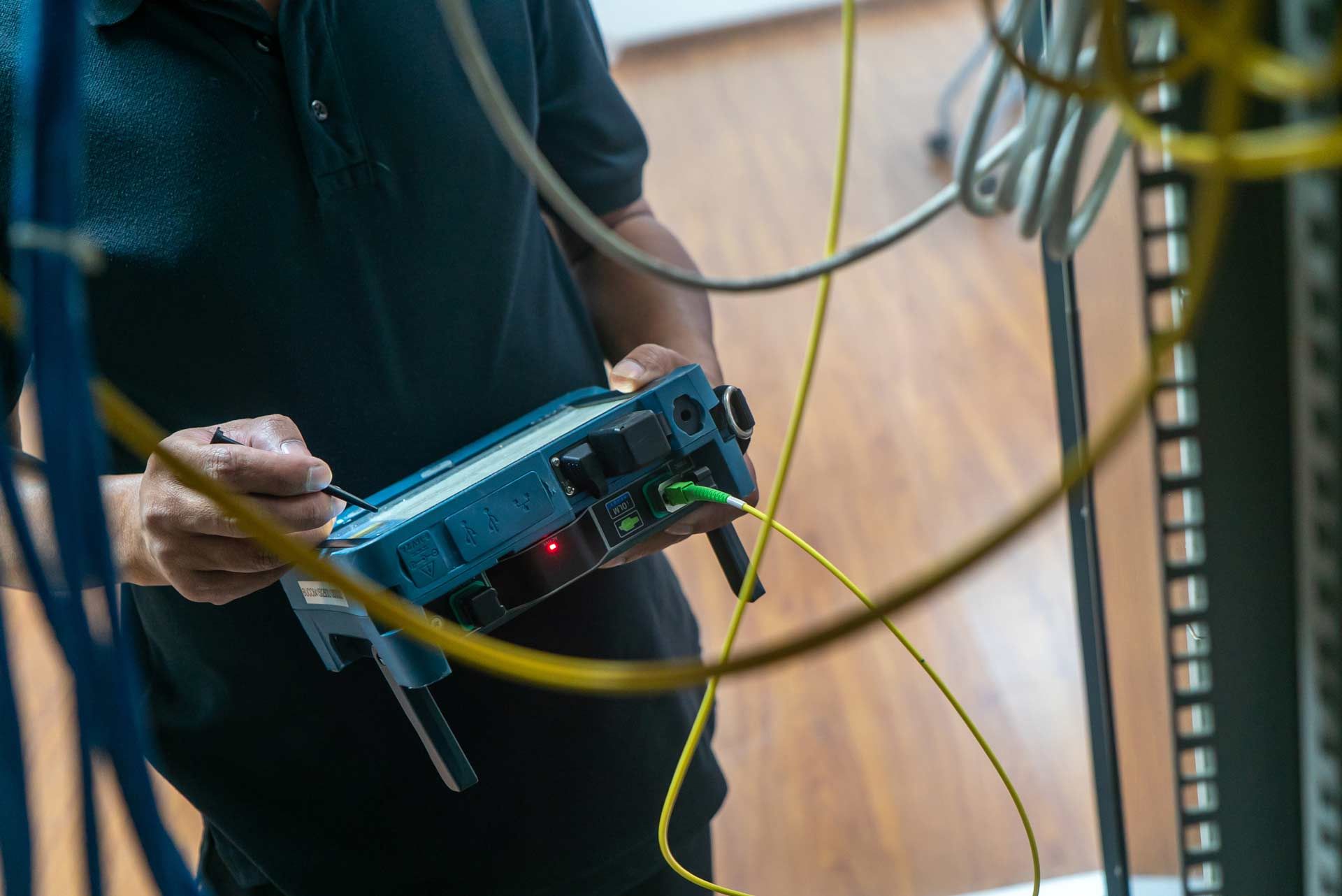 Person testing network cables with a blue testing device in a server room. Yellow and blue cables are visible.