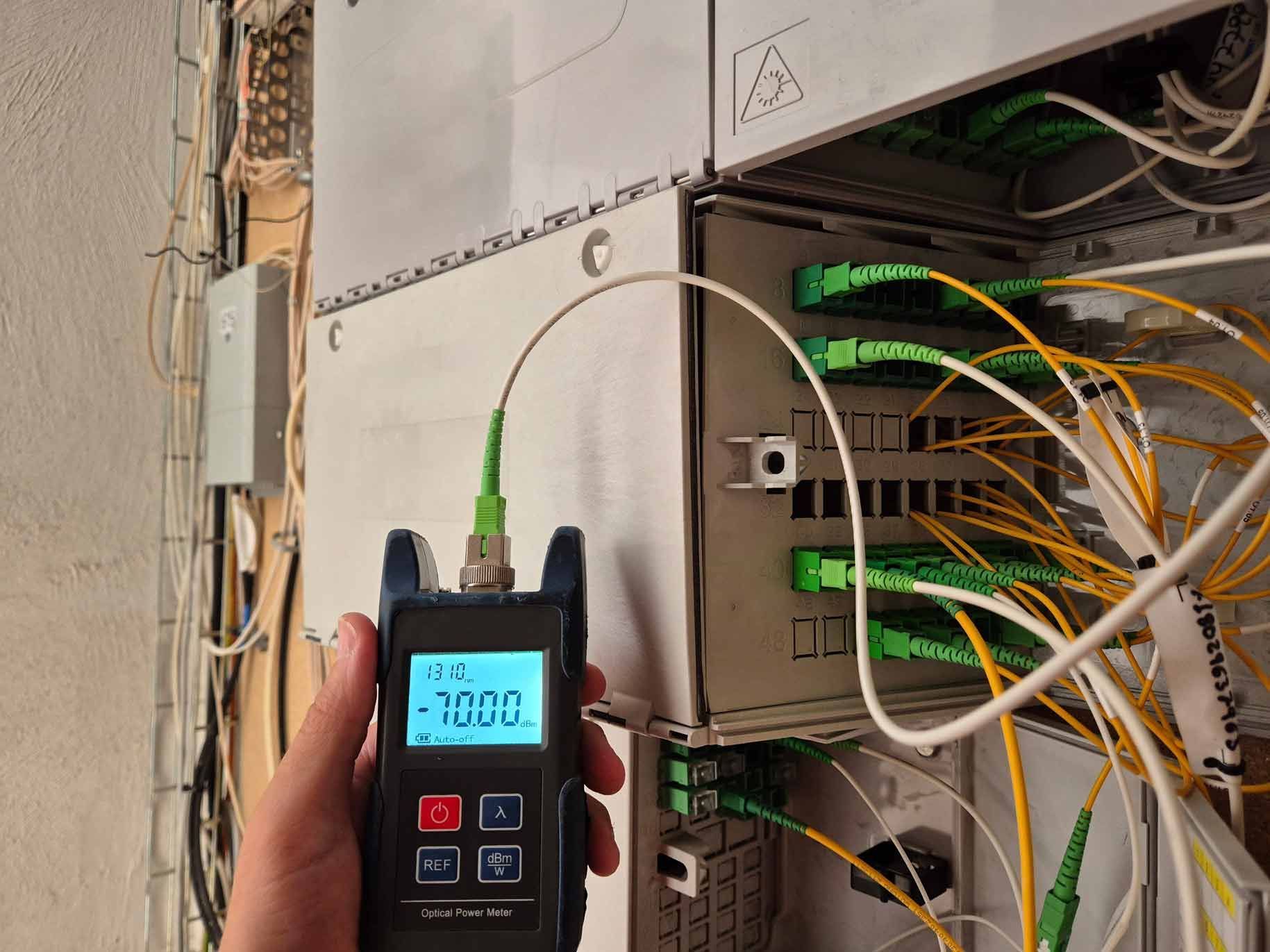 Hand holding a fiber optic meter connected to a white cable and network equipment with multiple green cables.