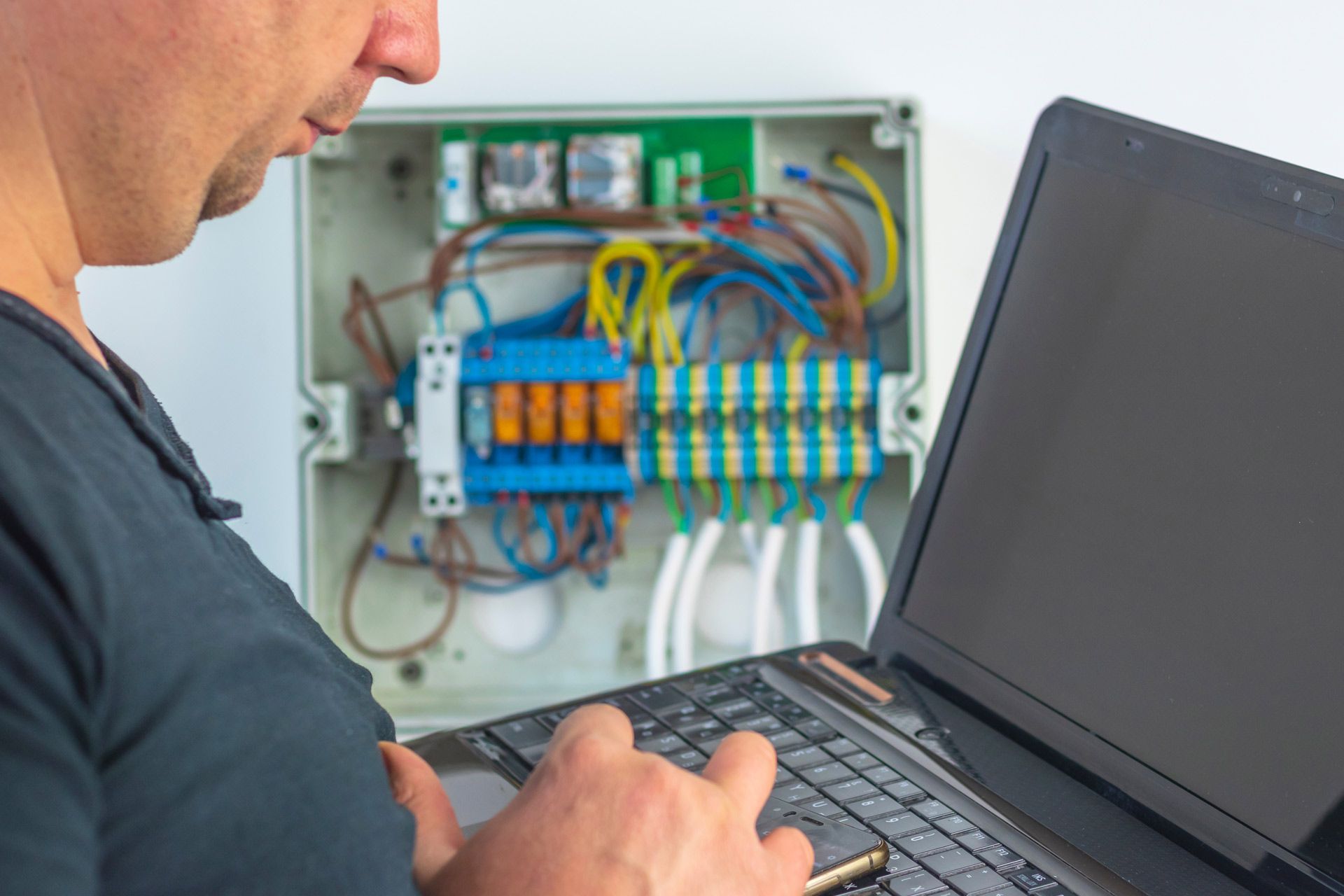 Person working on electrical panel, using laptop, white enclosure, wires, and circuitry visible.