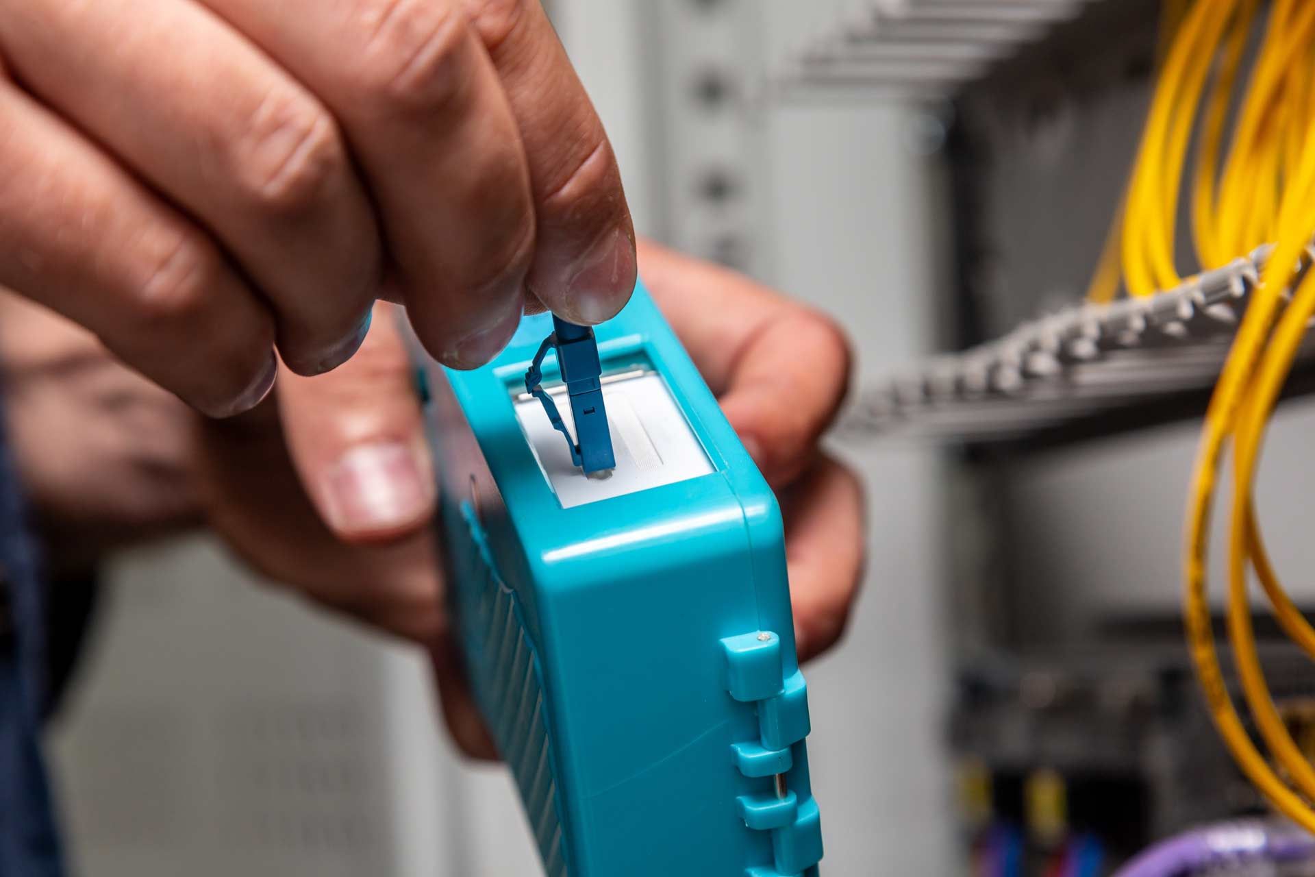 Person inserting a blue fiber optic cable connector into a turquoise testing device in a server room.