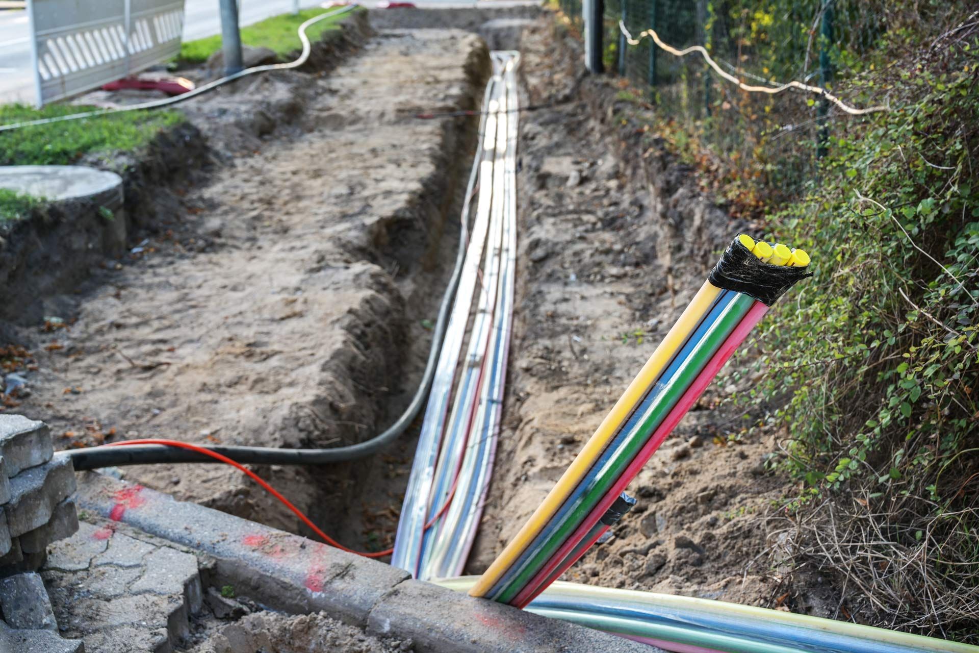 Trench with multiple cables laid in it, likely for utilities. Colors: silver, black, yellow, green. Outdoors setting.