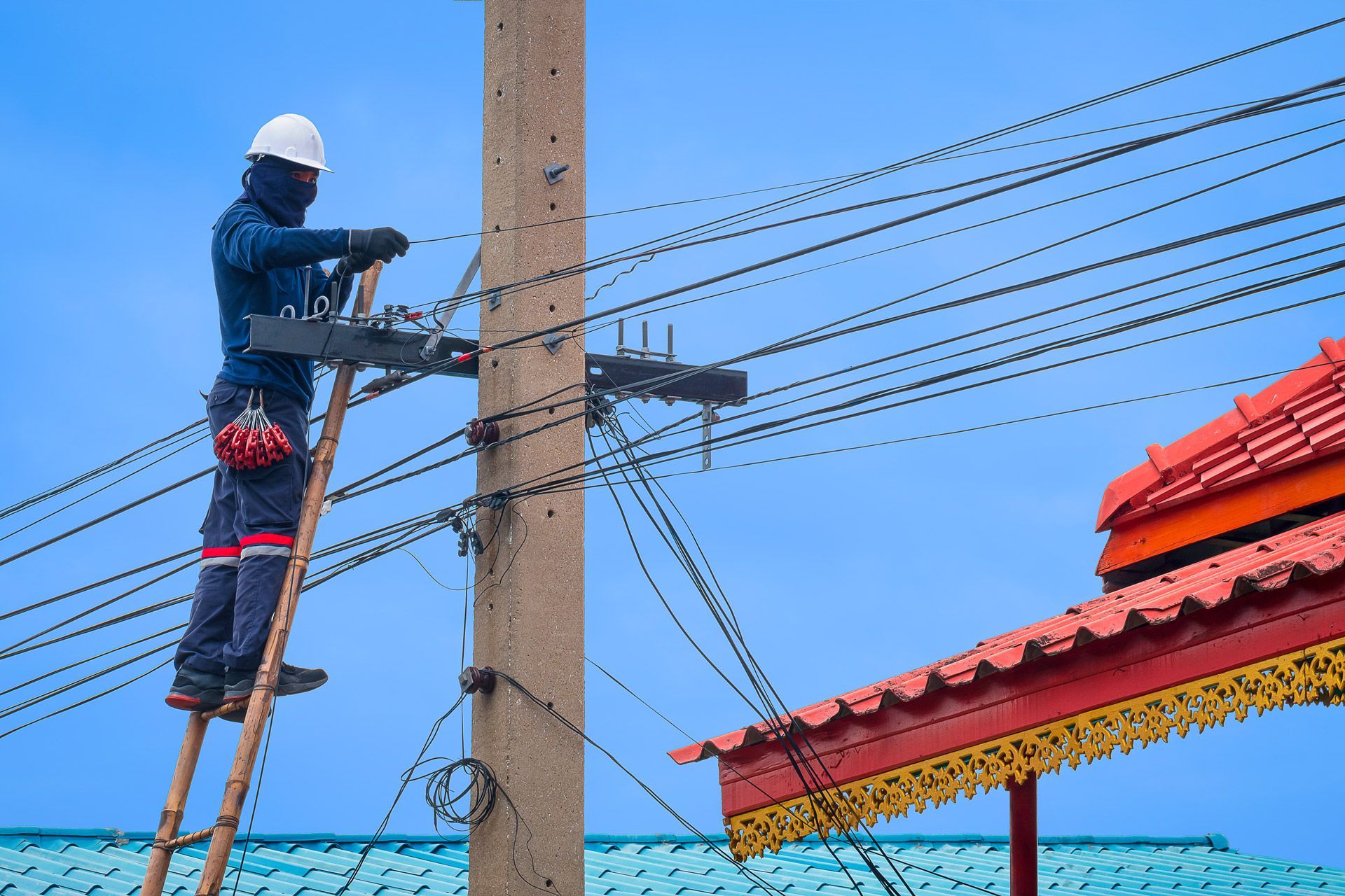 Electrician on a ladder working on power lines attached to a pole. Blue sky, red roof nearby.