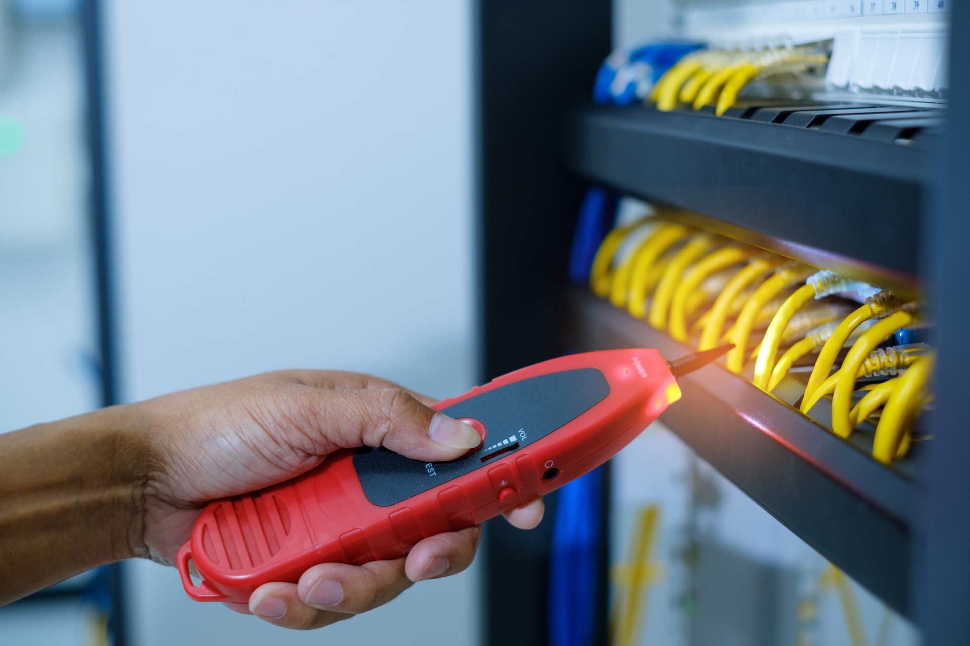 Hand holding a red network cable tester, probing yellow cables in a server rack.
