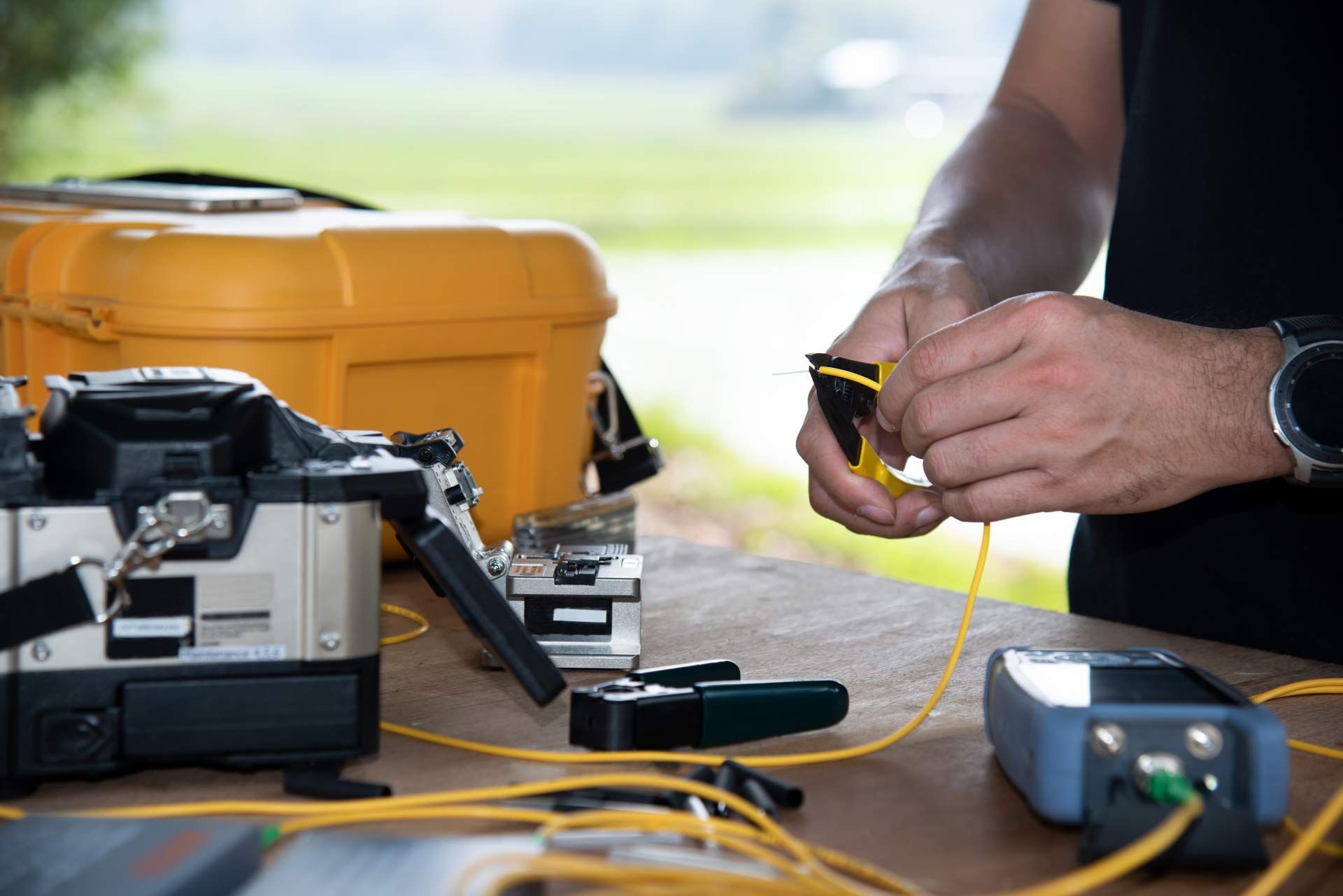 Person working with fiber optic cables outdoors, using tools on a table with equipment.