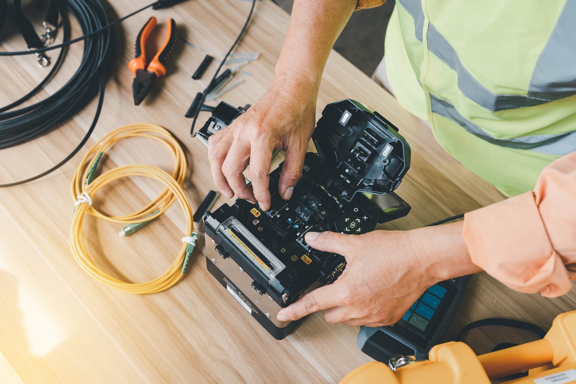 Person in safety vest working on black camera equipment on a wooden table with tools and cables.