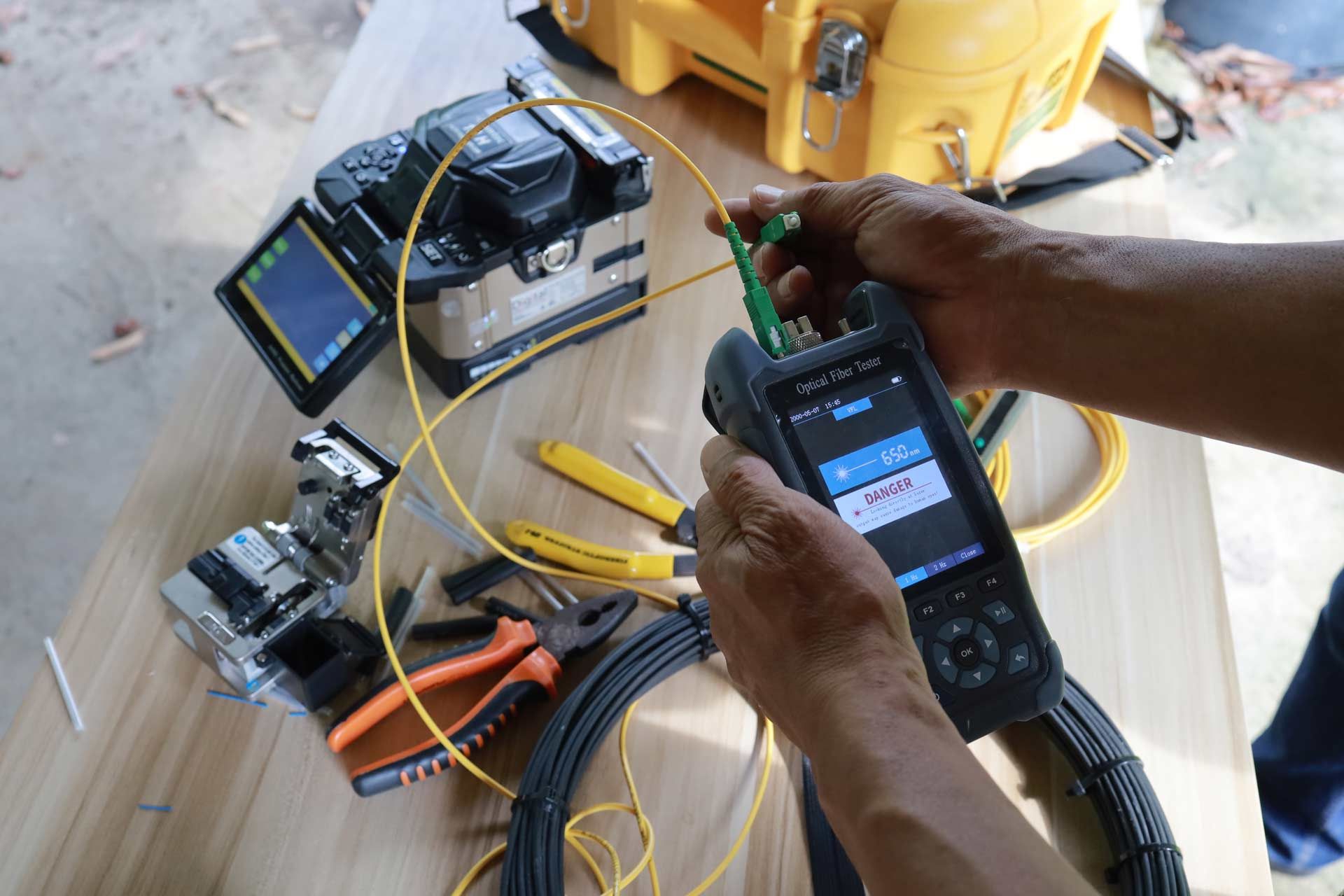 A person testing a fiber optic cable with a handheld device, surrounded by equipment on a wooden surface.