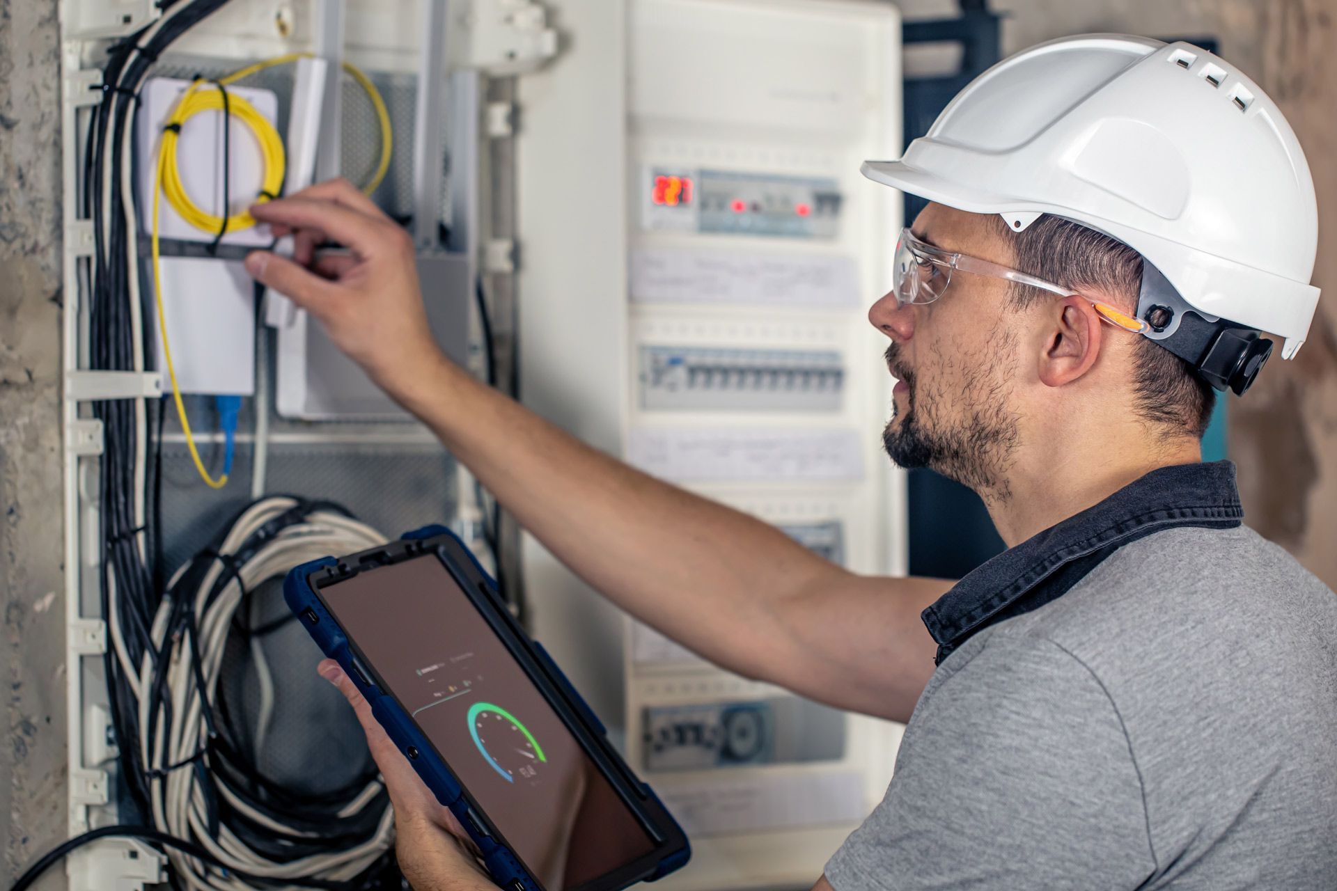 Electrician in a hard hat inspecting electrical panel, holding a tablet with data readings.