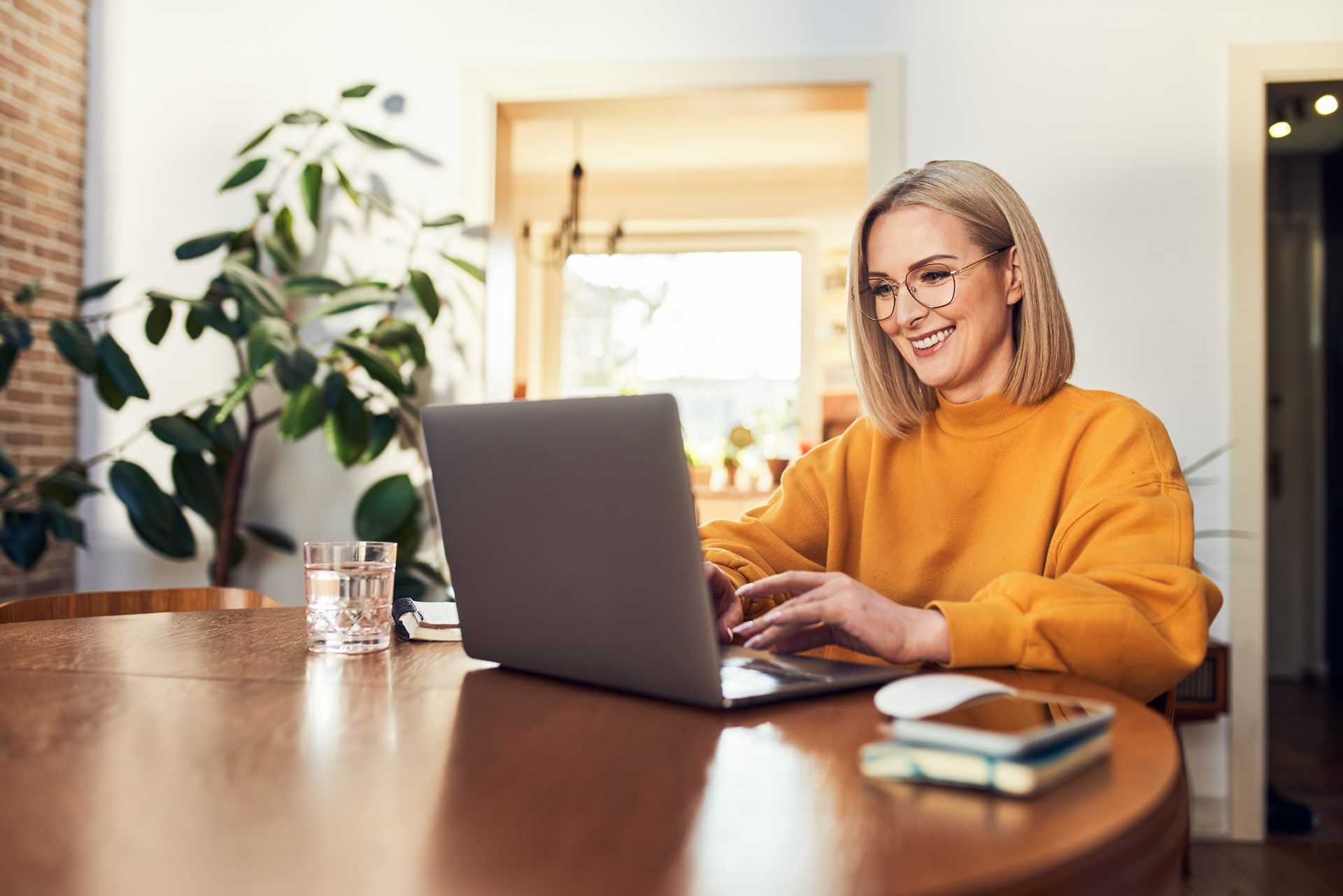 Woman in yellow sweater using laptop at a wooden table, smiling indoors.