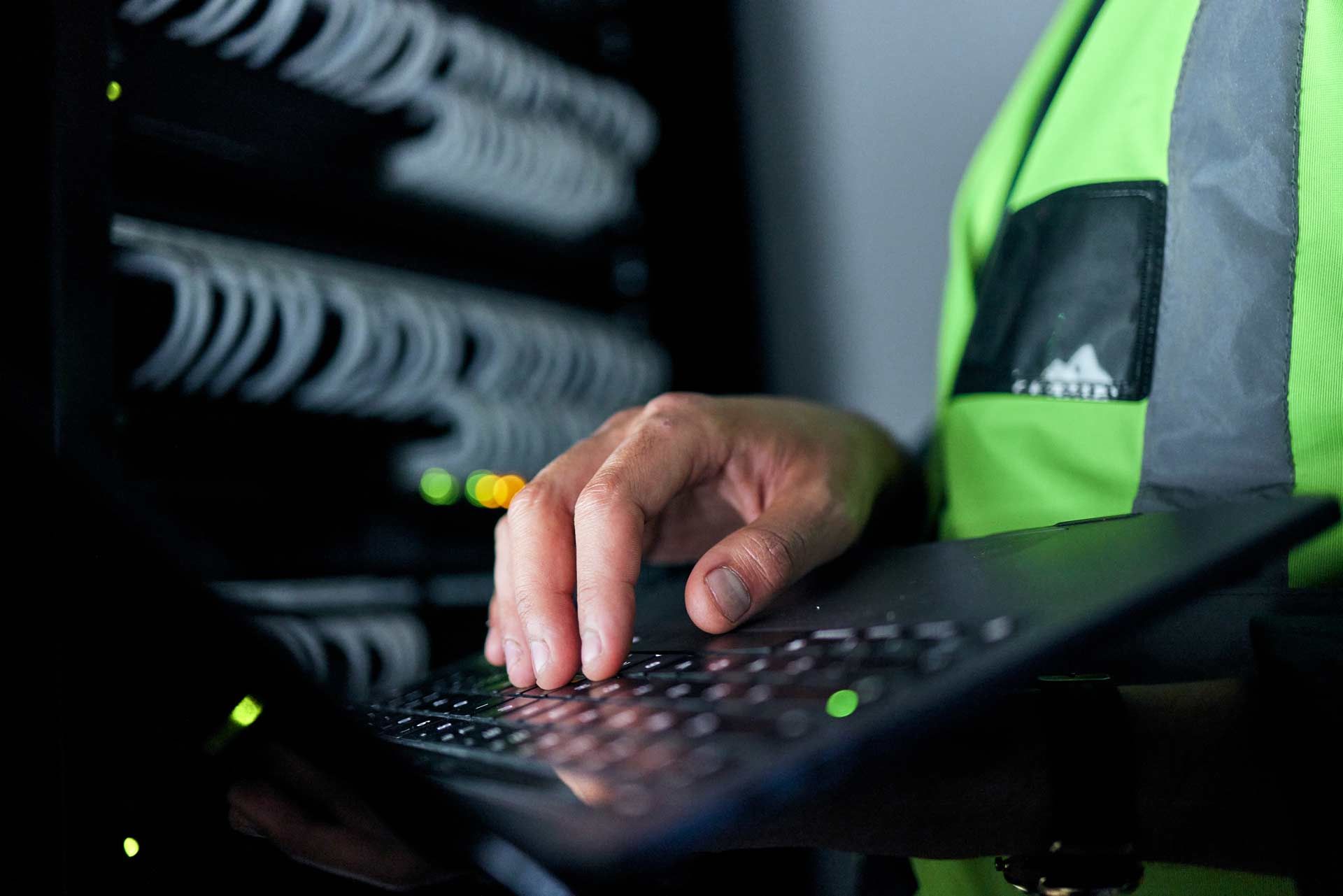 Person in safety vest using a laptop in a server room, typing with fingers.