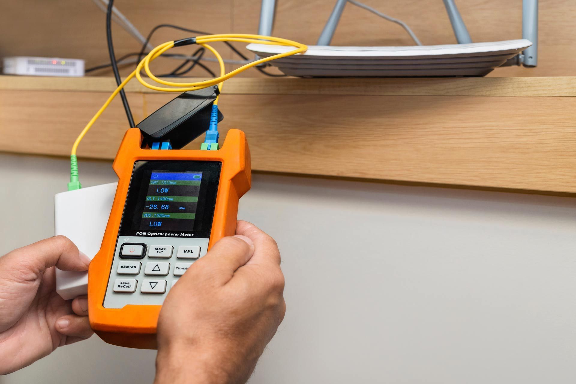 Person holding a fiber optic testing device, connected to a router and a wall-mounted box.