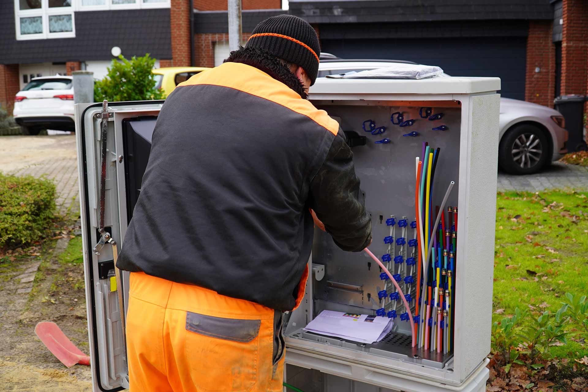 Worker in orange vest and pants working on a gray utility box outside.