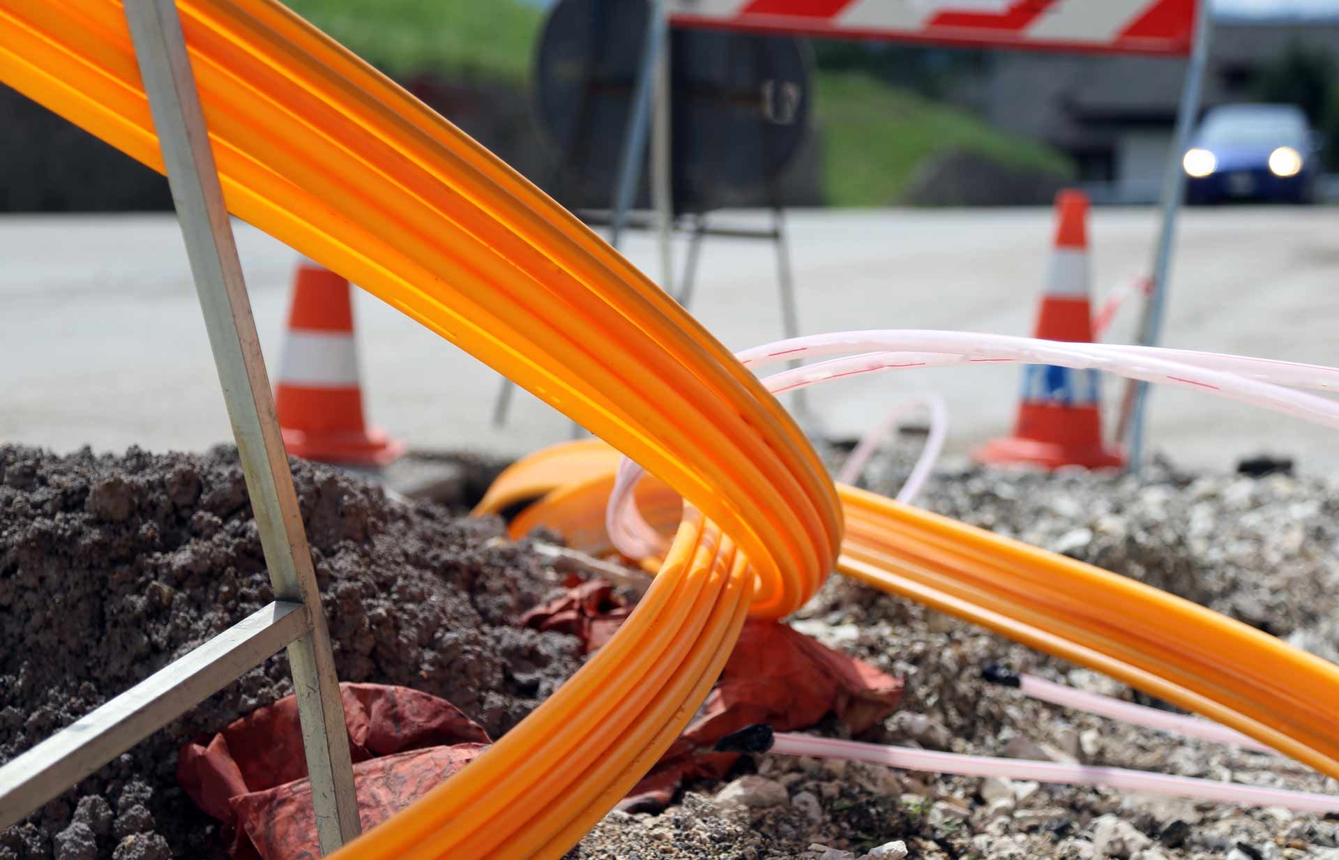 Orange fiber optic cables on gravel at a construction site, traffic cones in background.