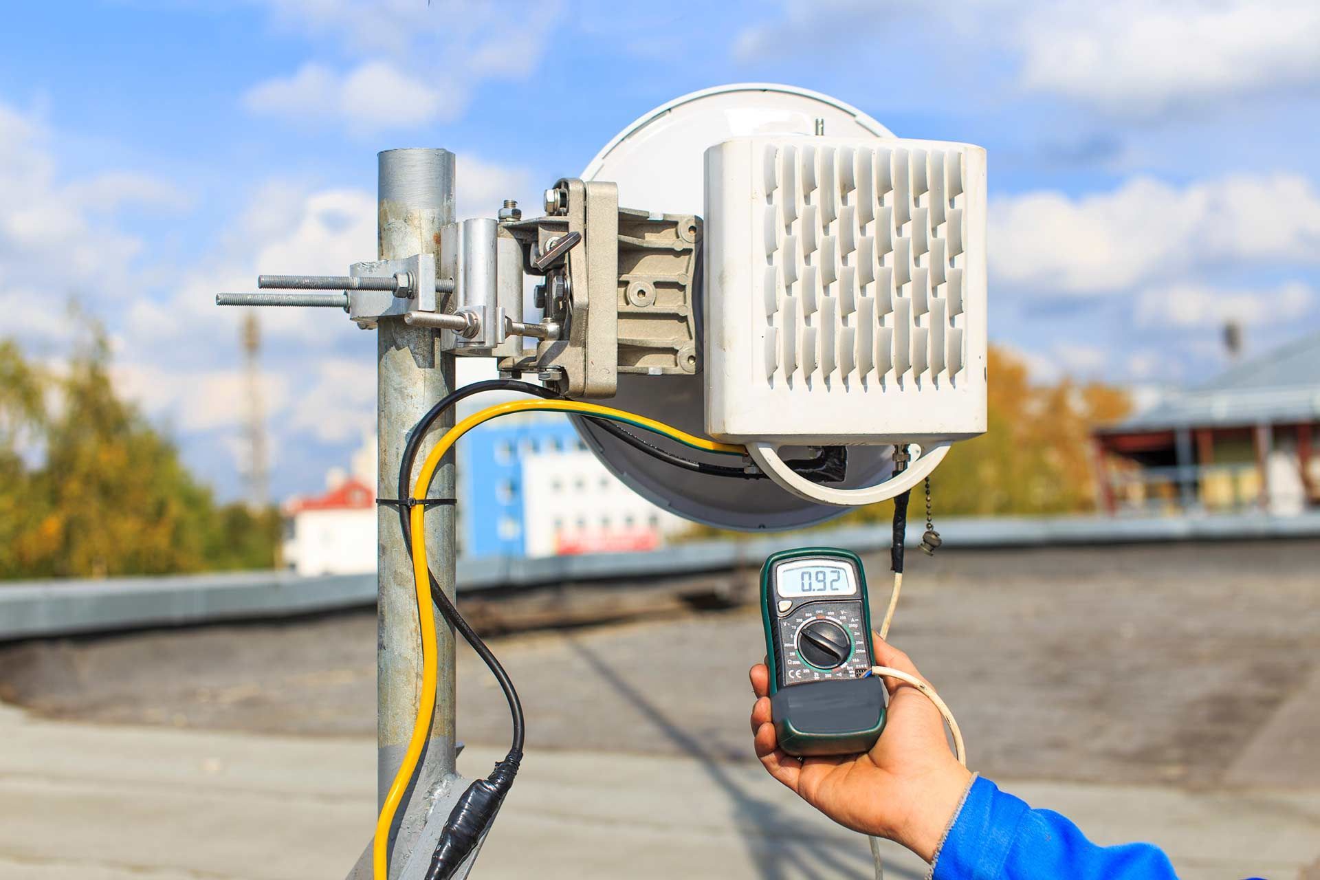 Person testing a cellular antenna on a rooftop with a multimeter; cables attached.