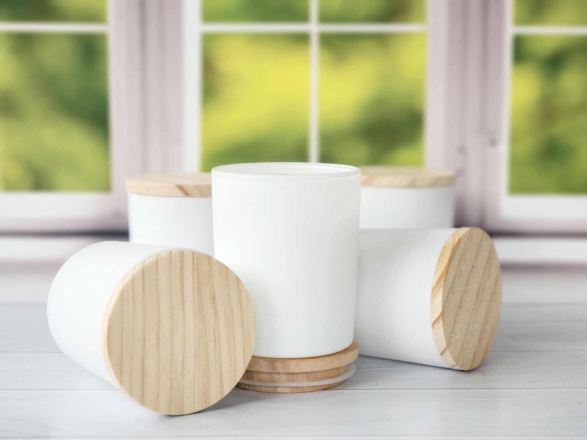 Four white cylindrical containers with wooden lids and bases on a white surface, with a blurred window background.