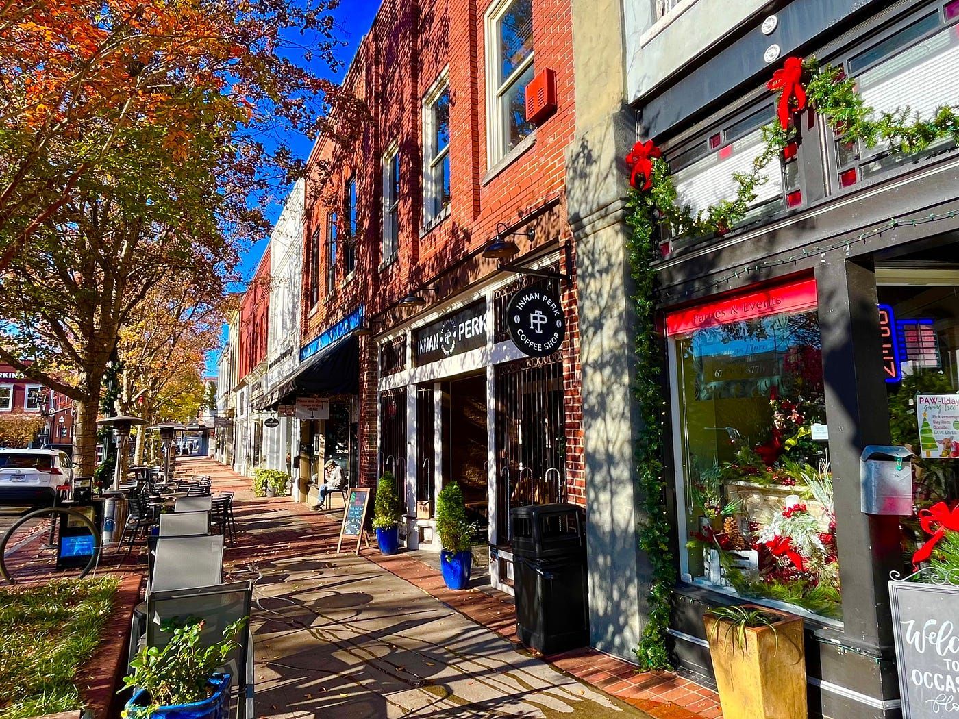 A sunny street scene in a historic town featuring red brick storefronts, autumn trees, and outdoor sidewalk seating.