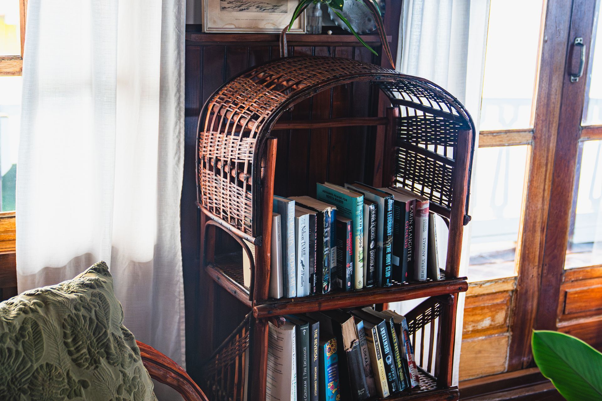A wicker bookshelf filled with books in a living room next to a chair.