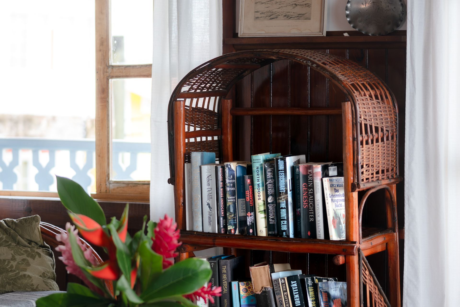 A wicker bookshelf filled with books and flowers in front of a window.