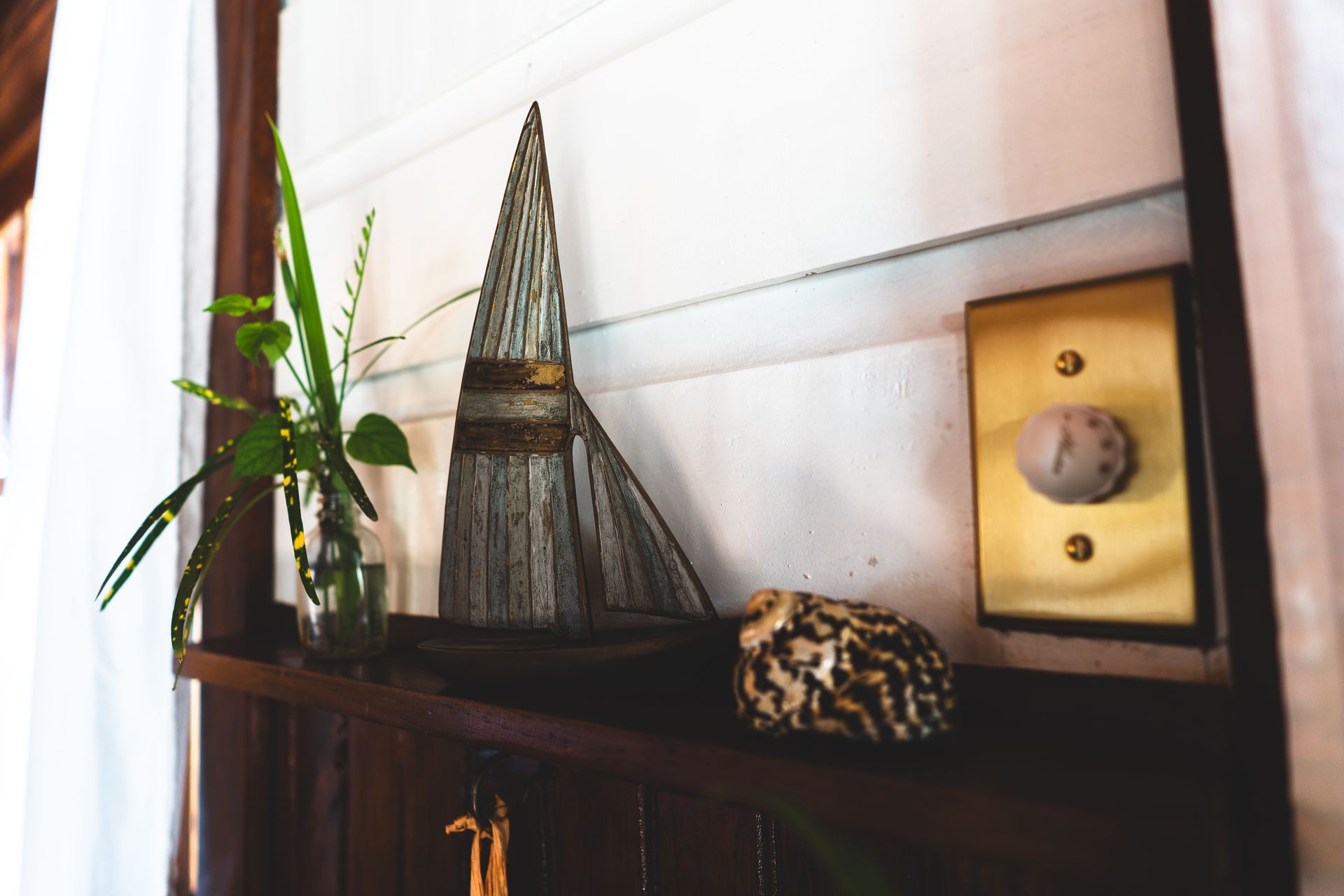 A wooden boat, flowers, and a seashell  are sitting on a shelf next to a light switch.