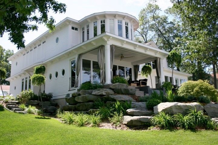 White house with multiple levels, porch, and a rocky landscaping. Lush green lawn and trees surround the home.