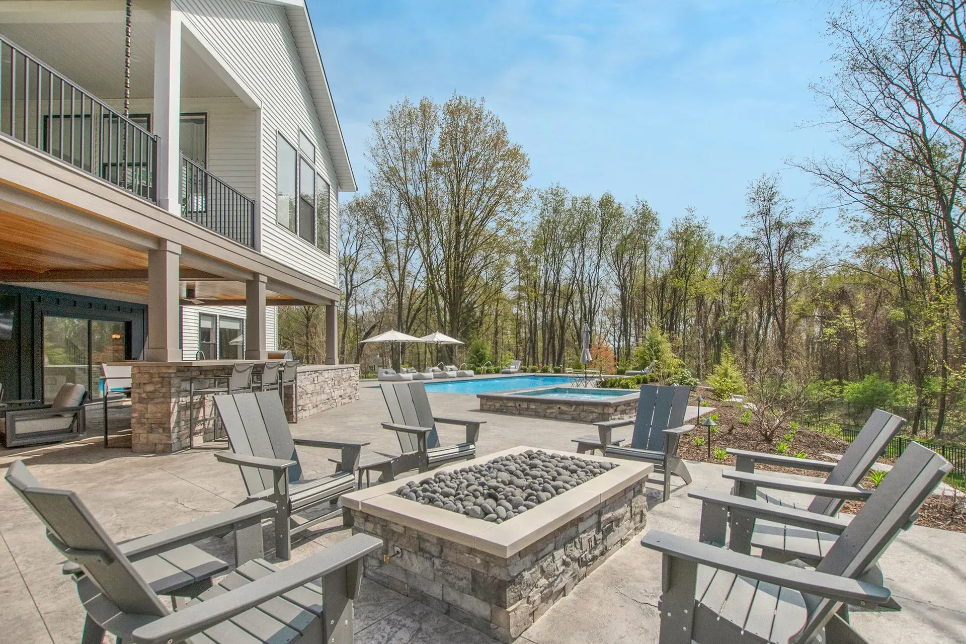 Patio with fire pit and Adirondack chairs, house with balcony, pool in the background. Sunny day.
