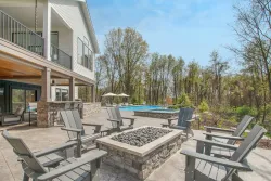 Patio with fire pit and Adirondack chairs, house with balcony, pool in the background. Sunny day.
