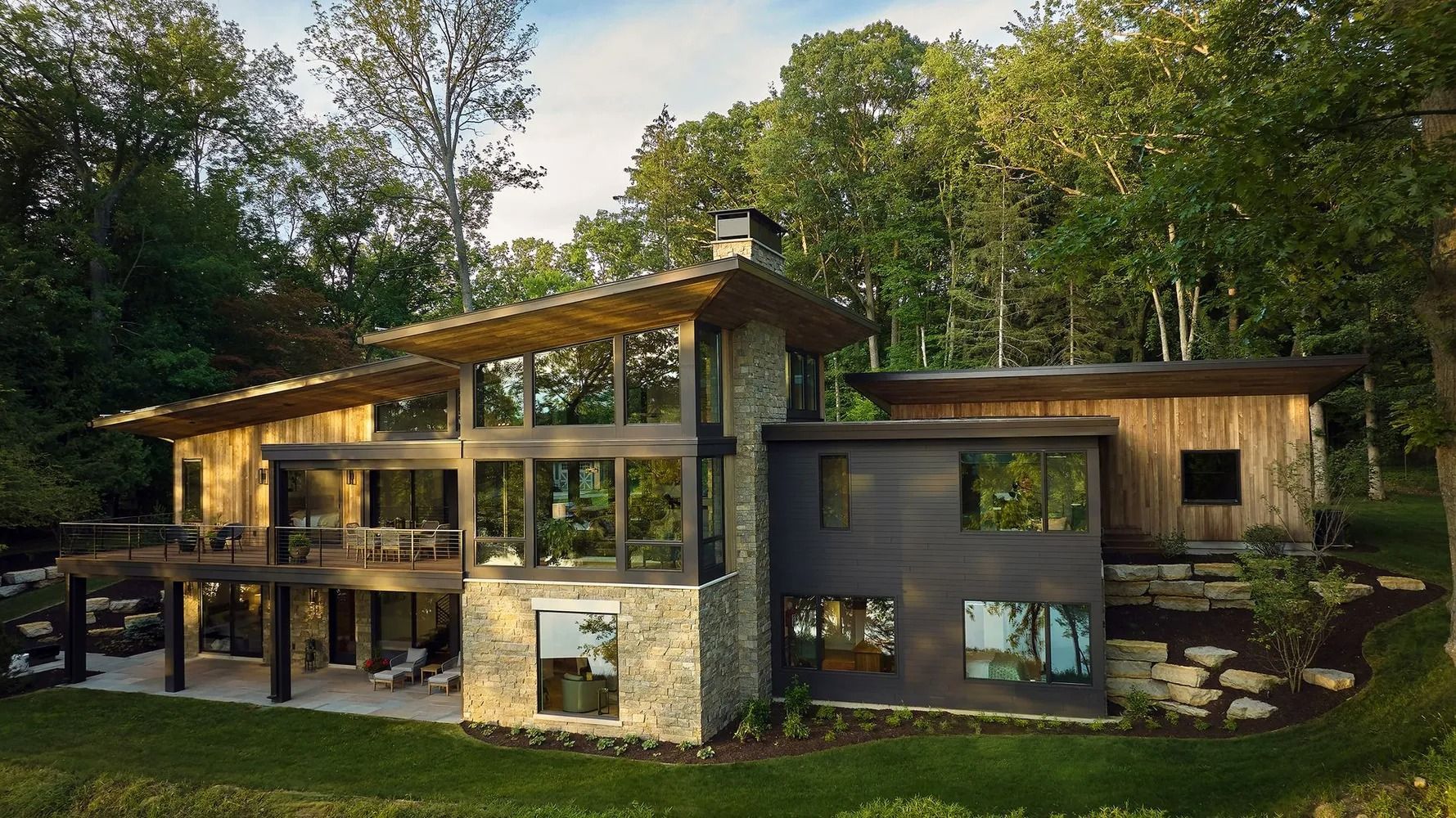 Modern house with stone and wood exterior, large windows, and a deck, set amidst trees.