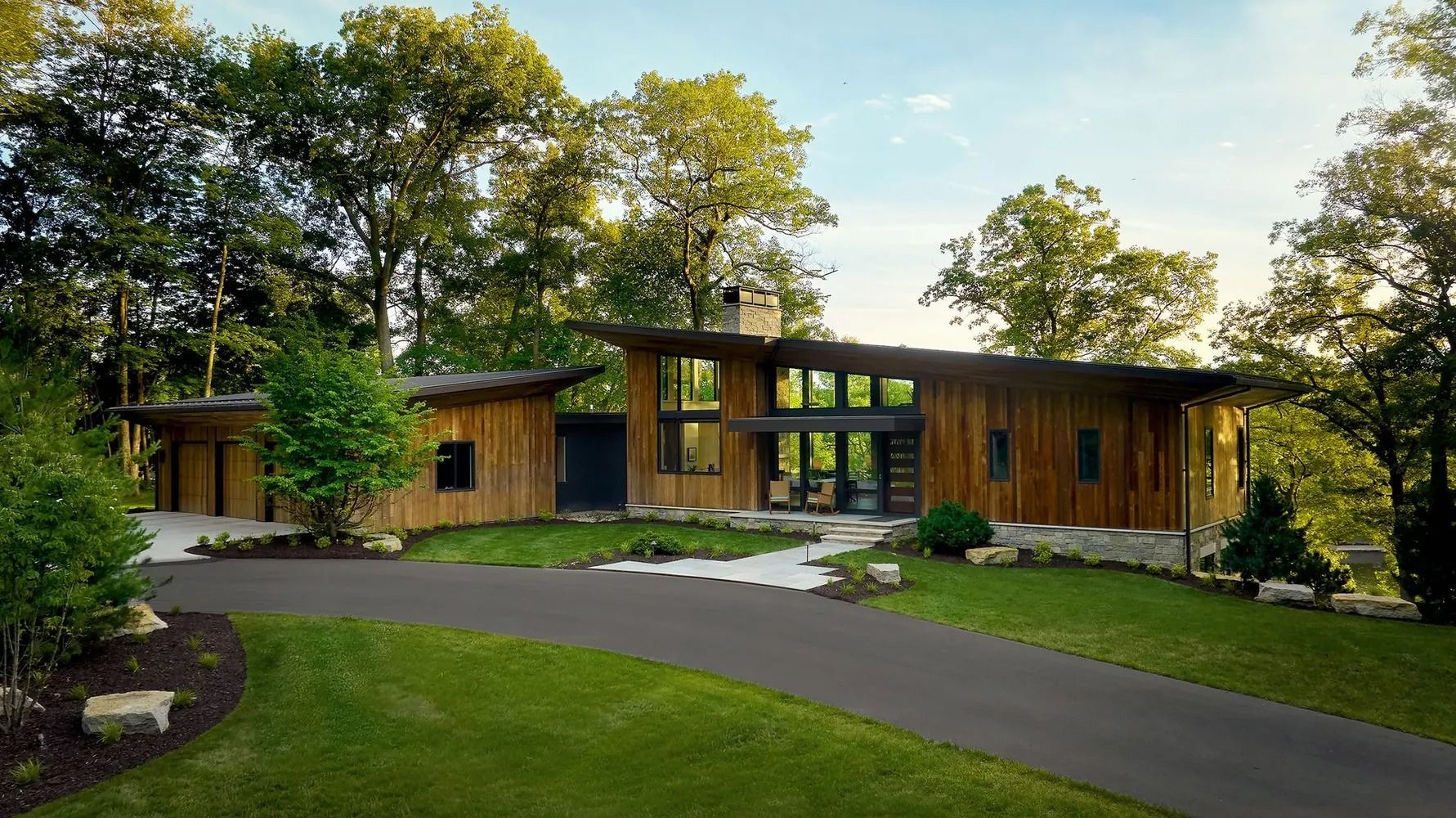Modern wood-paneled house with a sloping roof, surrounded by trees and a green lawn; a driveway leads up to it.