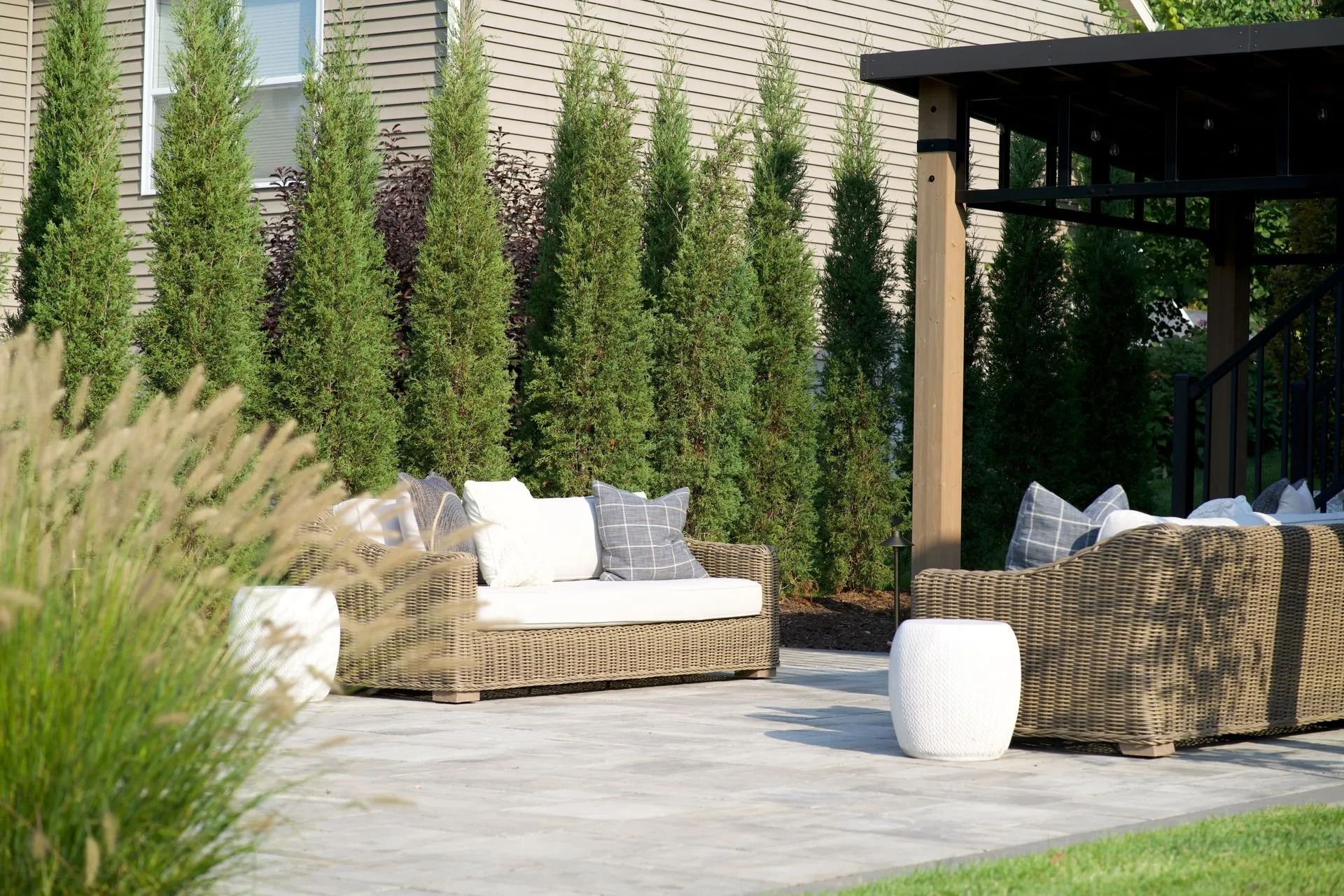 Outdoor seating area with wicker furniture on a stone patio, evergreen trees, and a pergola.
