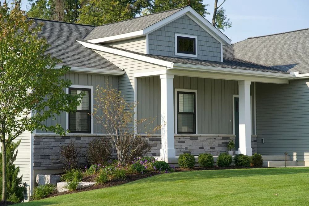 Gray house with stone base, front porch, and dormer window, set on a green lawn.